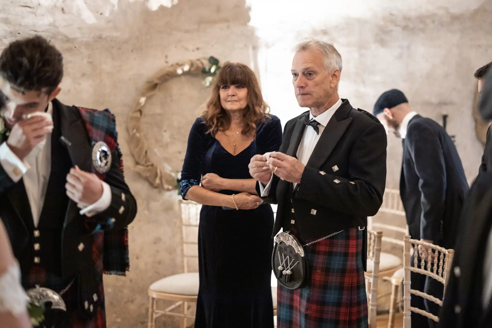 A woman and man, both in formal attire, stand in a room. The man wears a kilt and sporran, while the woman is in a dark dress. Others in similar attire are partially visible in the background.