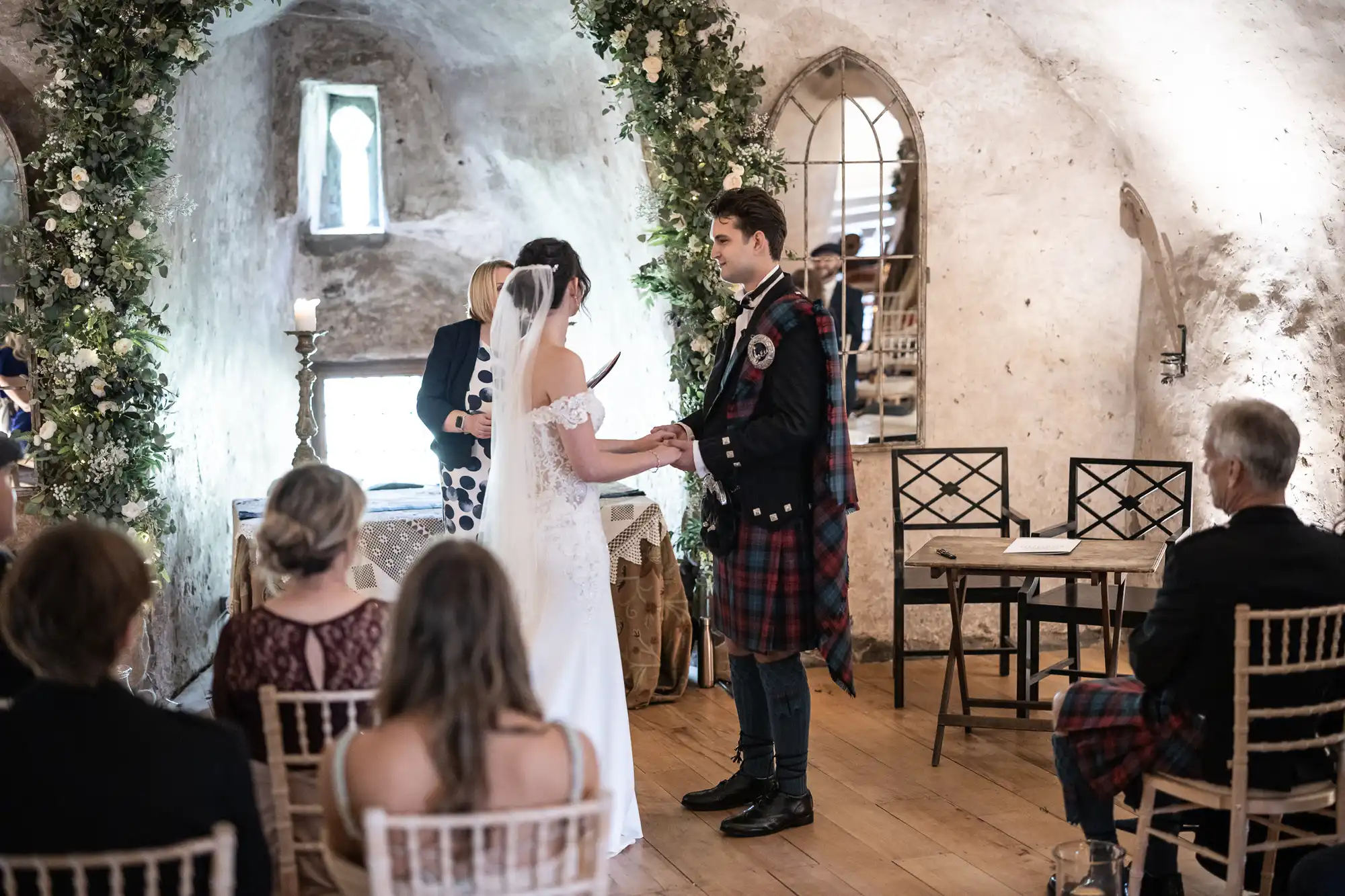 A bride and groom stand facing each other during a wedding ceremony in a rustic, stone-walled setting. Guests are seated, observing the couple. The groom wears a kilt, and the wedding arch is adorned with greenery.