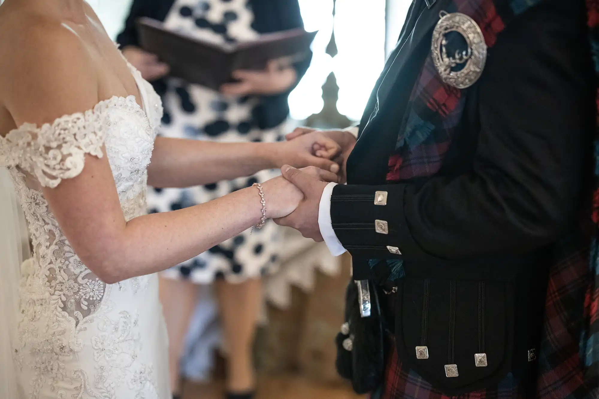 A bride and groom, with the groom wearing a traditional tartan outfit, hold hands during their wedding ceremony.