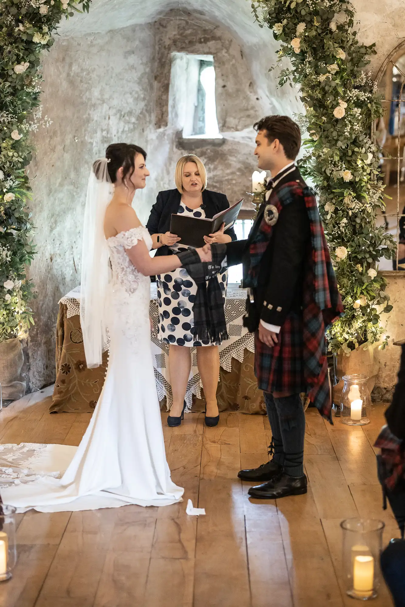 A couple stands facing each other exchanging vows in a decorated indoor venue, with a person officiating the ceremony.