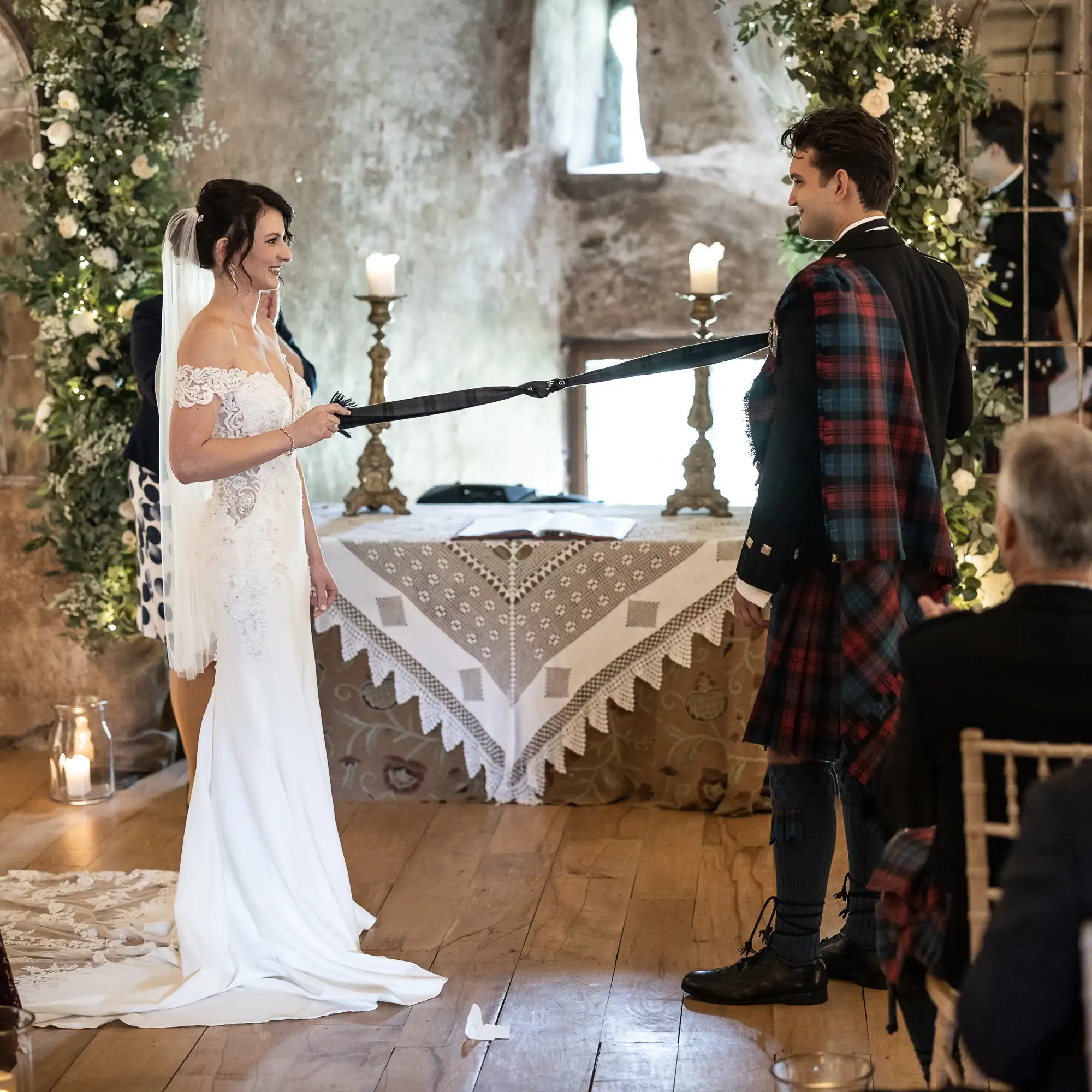 A bride and groom, dressed in a wedding dress and kilt respectively, stand facing each other, holding a tartan ribbon, in a room decorated with flowers and candles.