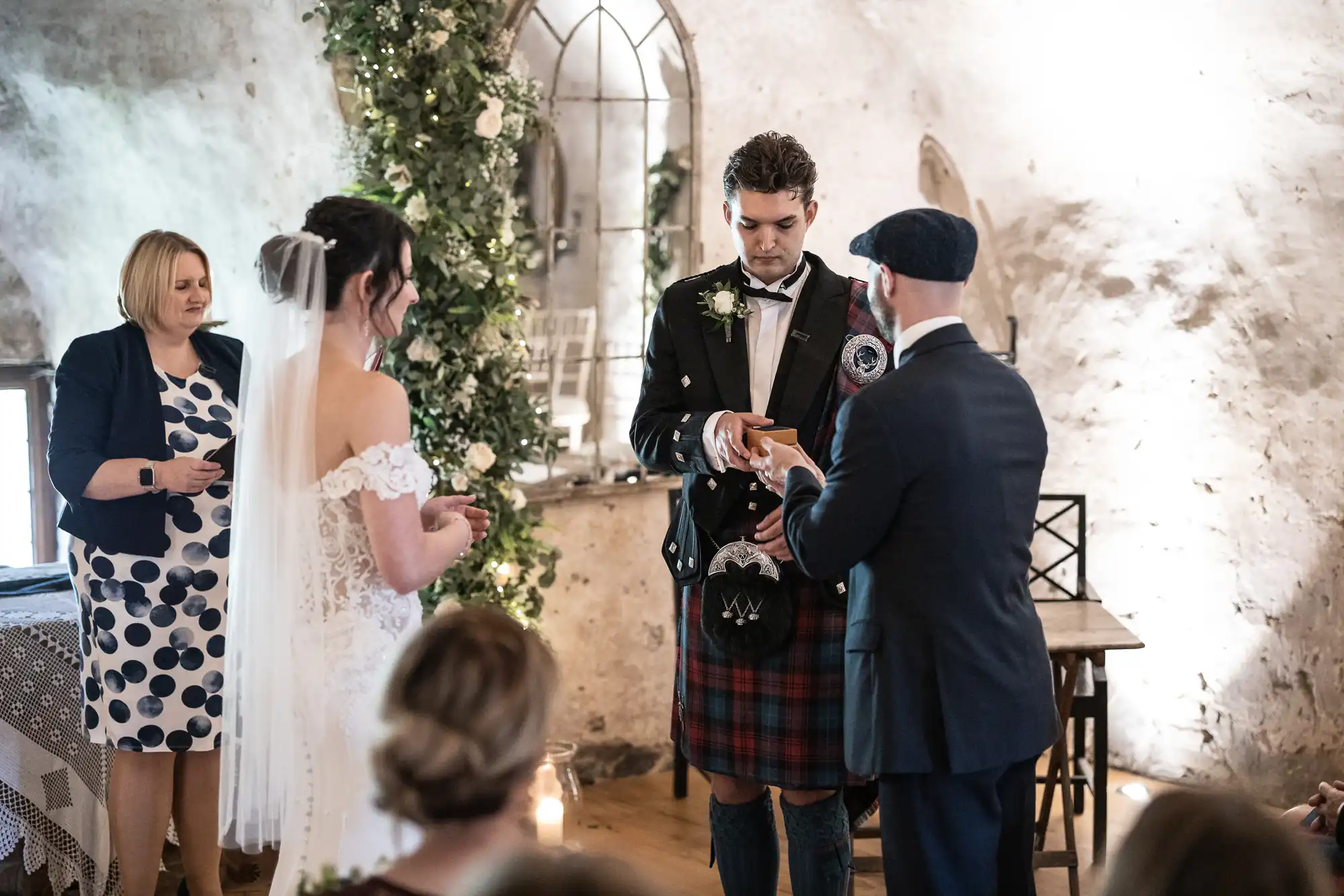 A couple stands in a rustic wedding ceremony exchanging rings, with a man in a kilt and a celebrant guiding them. Guests are seated in the foreground.