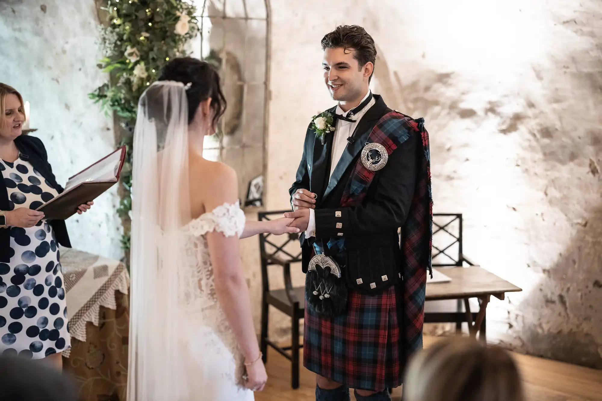 A couple stands facing each other during their wedding ceremony. The groom wears a traditional Scottish kilt, while the bride wears a white dress with a veil. An officiant stands beside them.