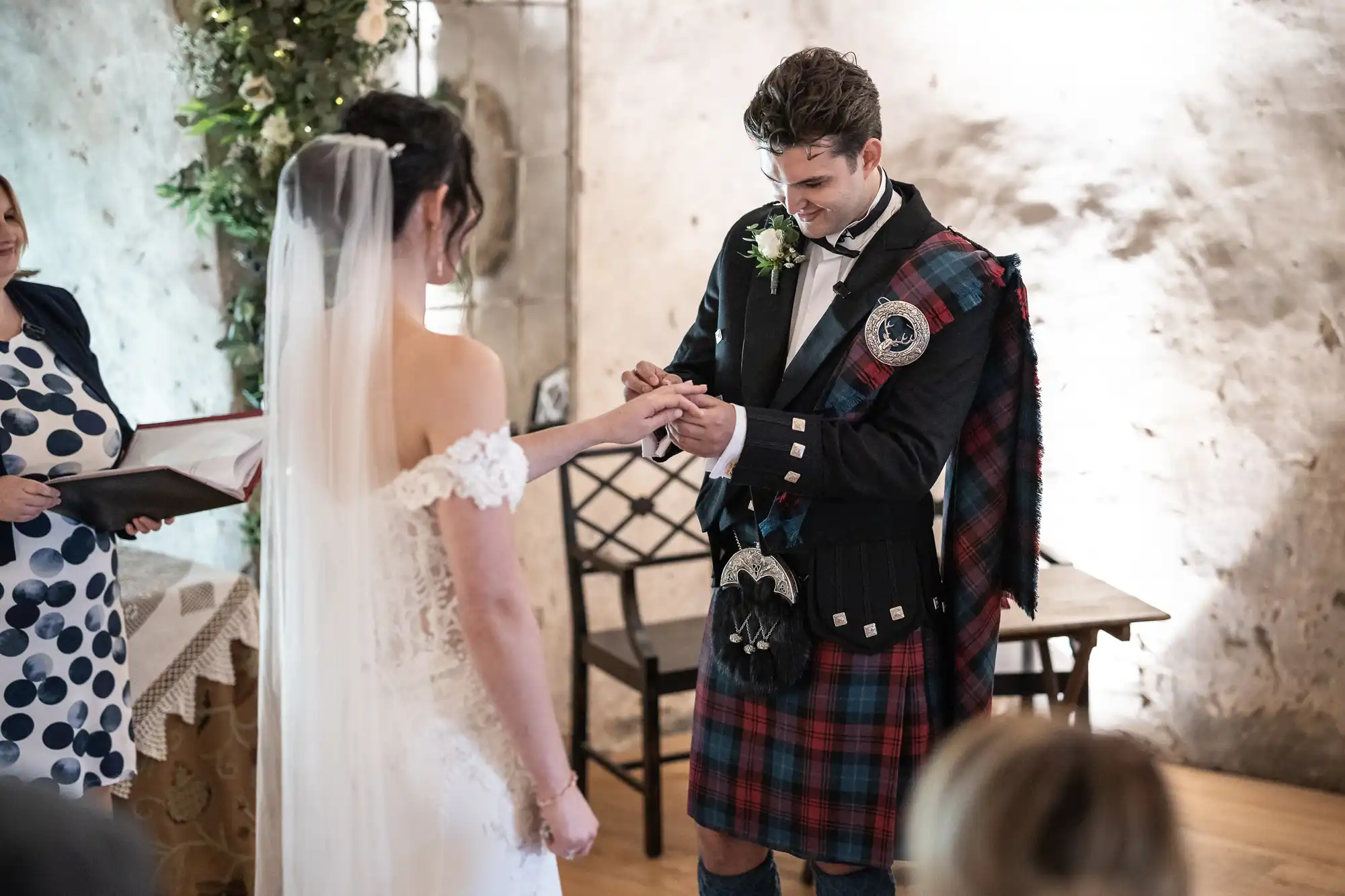 A bride and groom exchange rings during a wedding ceremony. The groom wears a traditional Scottish kilt outfit and the bride wears a white gown and veil. An officiant stands next to them holding a book.