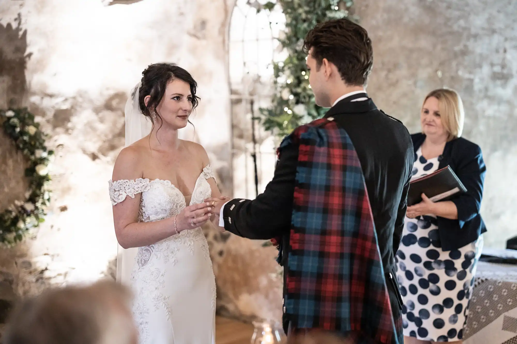 A couple in wedding attire is exchanging rings during a ceremony, with a woman holding a book standing beside them in a decorated, rustic setting.