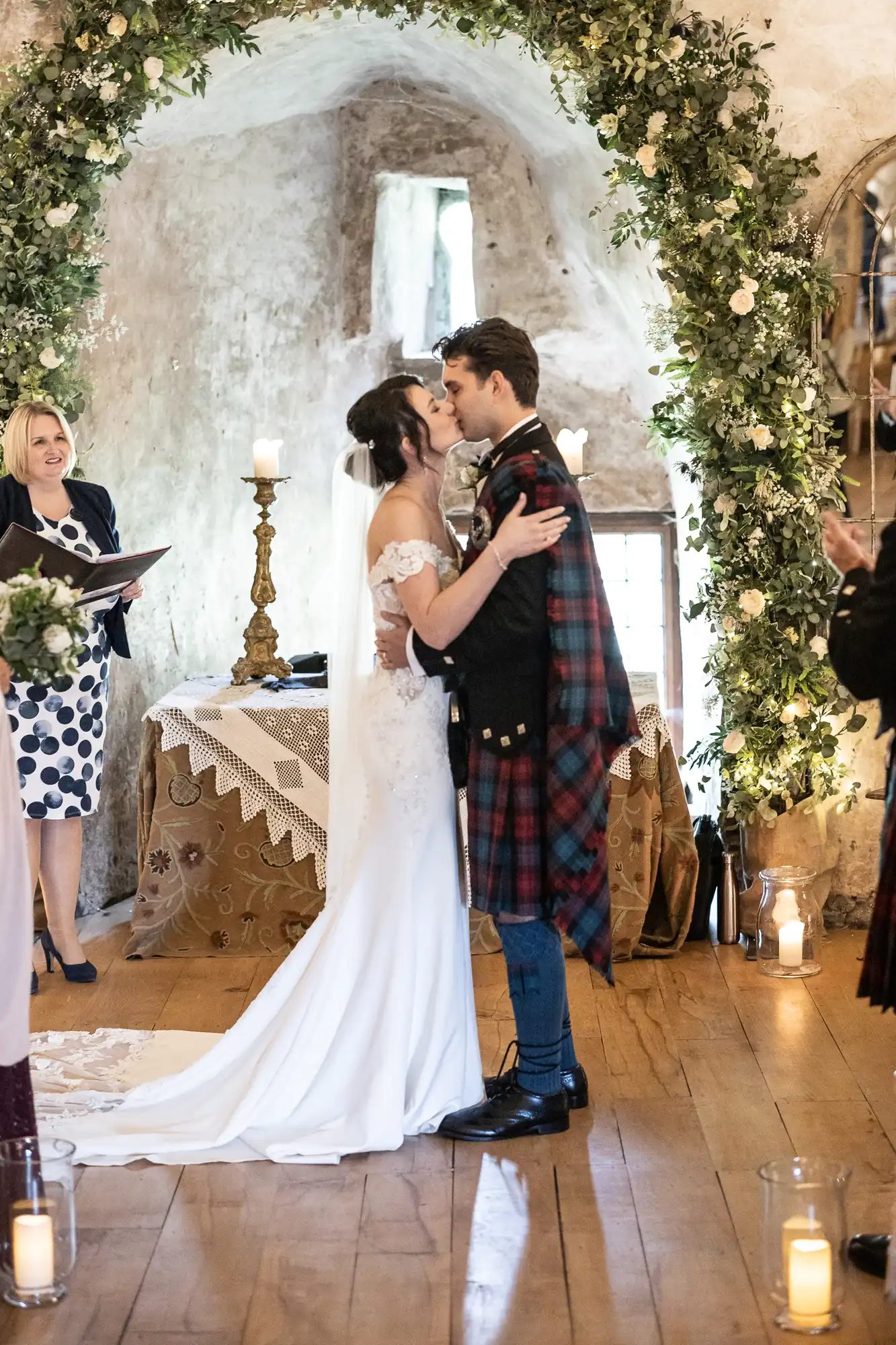 A wedding couple shares a kiss under an arch adorned with flowers, with candles and greenery in the background. The bride is in a white dress; the groom wears a kilt. An officiant stands nearby.