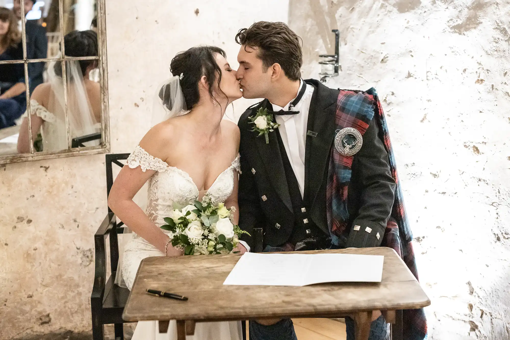 A bride and groom share a kiss while seated at a table with a pen and signed document. The bride holds a bouquet, and the groom wears a traditional Scottish outfit with a kilt and sash.