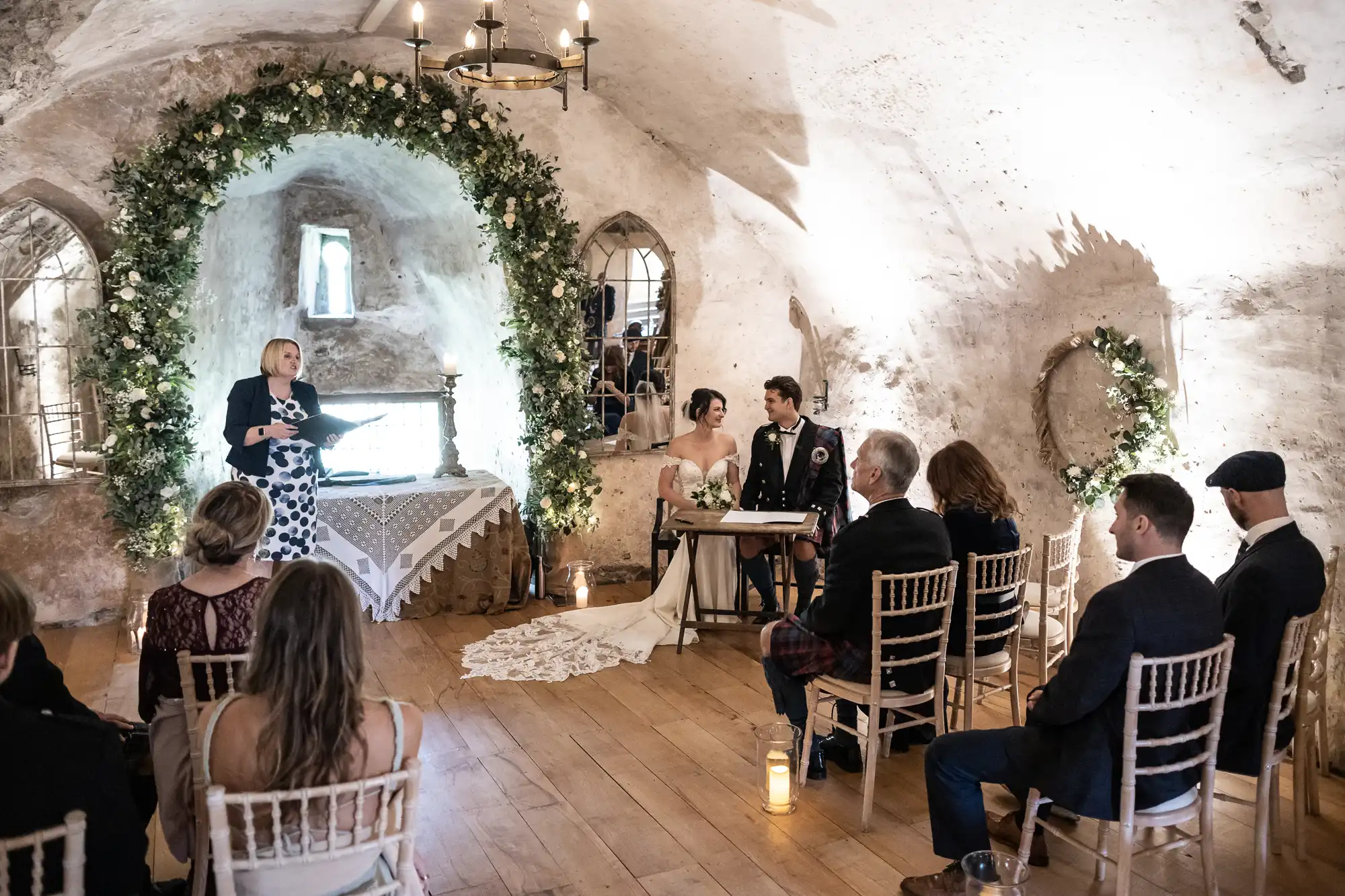 A couple sits at a table during their wedding ceremony in a rustic, stone-walled room with a floral arch. Guests are seated, and a woman stands to the side, speaking.