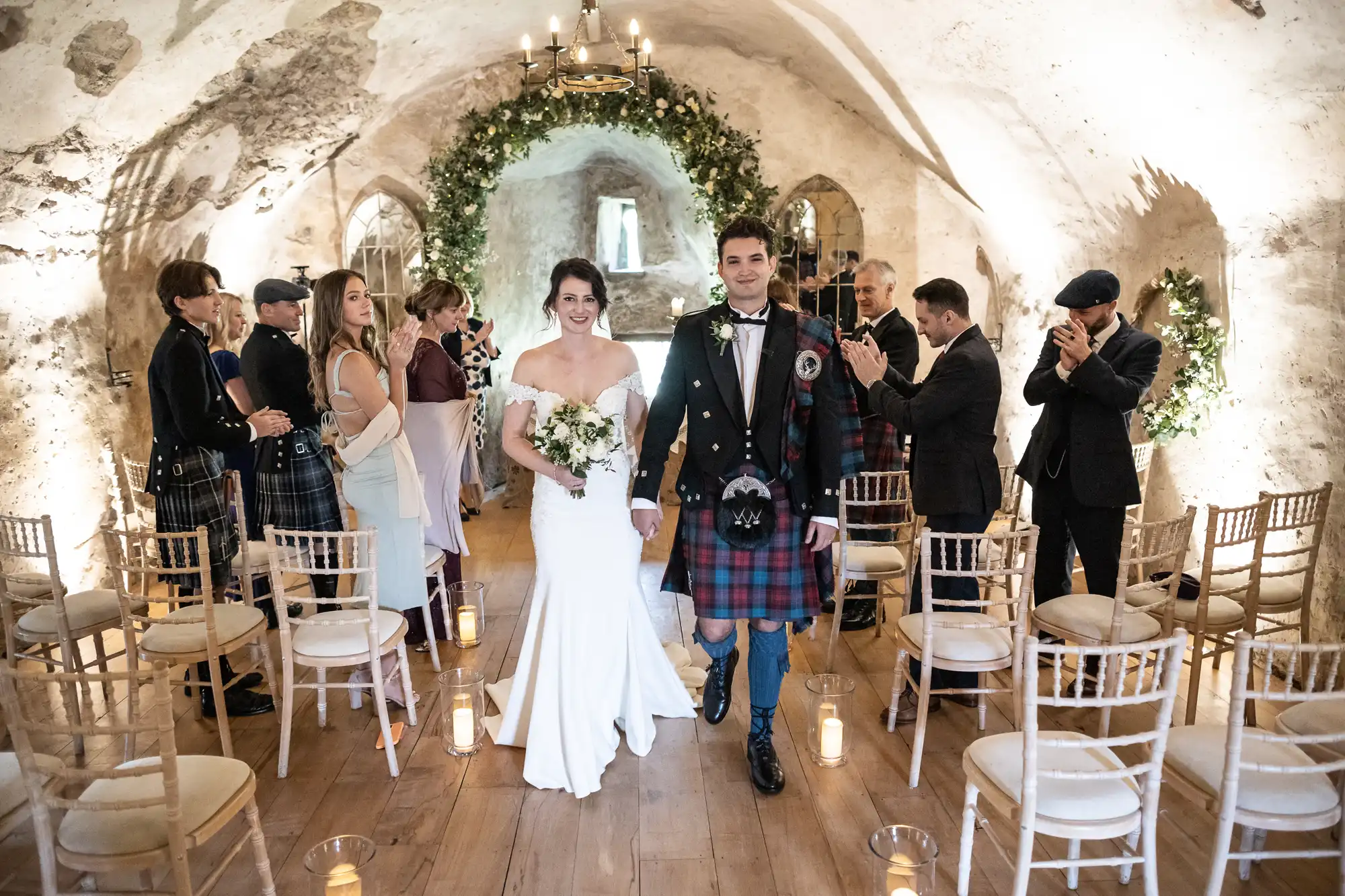 A bride in a white dress and a groom in a tartan kilt walk down the aisle of a decorated, rustic hall, followed by clapping guests.