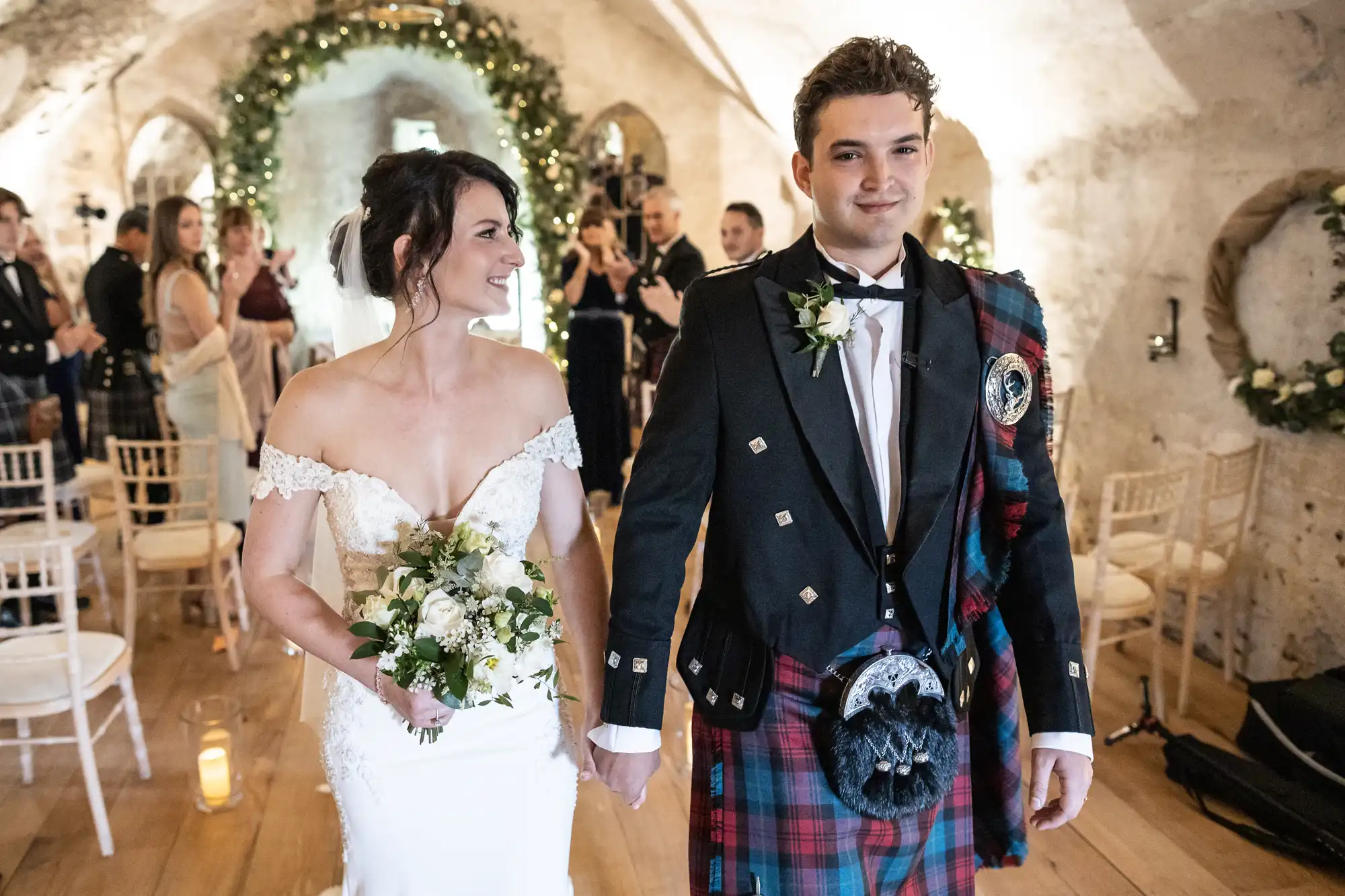 A bride and groom walk down the aisle in a rustic, decorated venue. The bride is in a white dress, and the groom is in traditional Scottish attire, including a kilt. Guests are visible in the background.