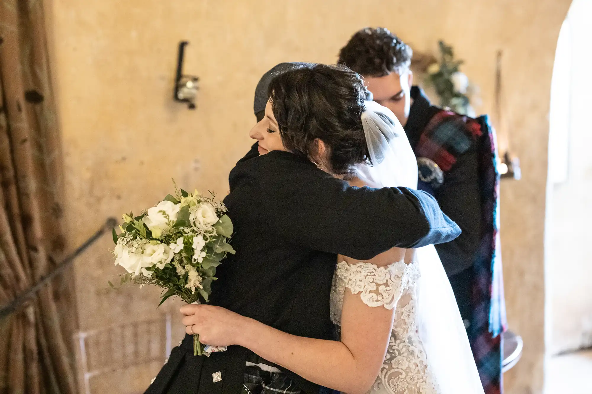 A bride in a white gown and veil hugs a man in dark attire holding a bouquet of flowers. Another person in formal wear stands in the background.