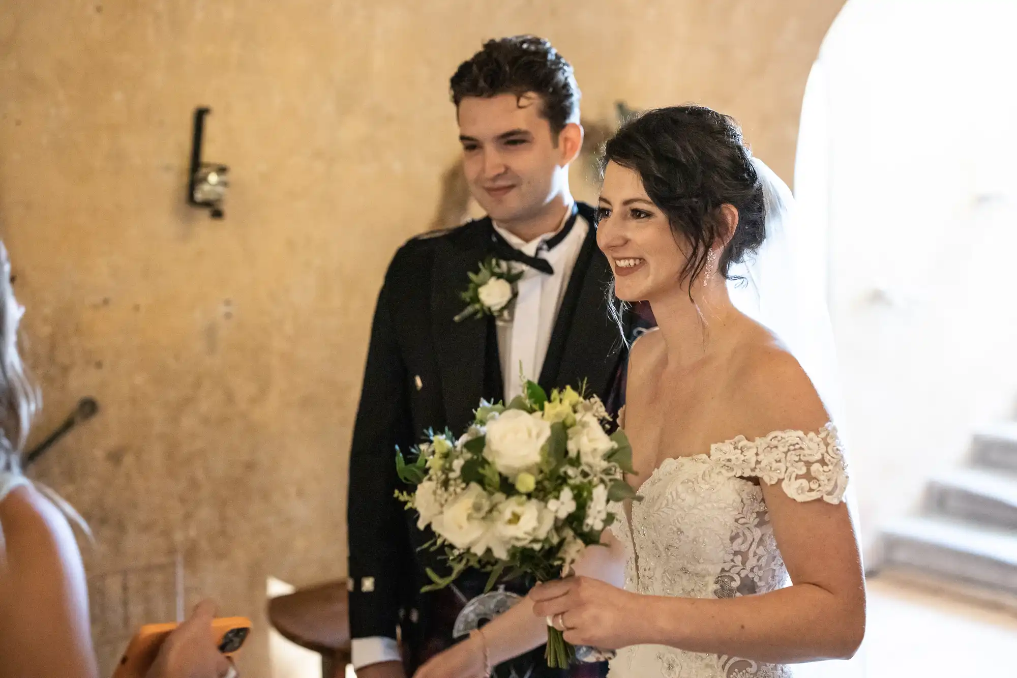 A bride in a white gown and veil holds a bouquet. A groom in a dark suit stands beside her. Both are smiling and facing someone not fully visible in the frame.