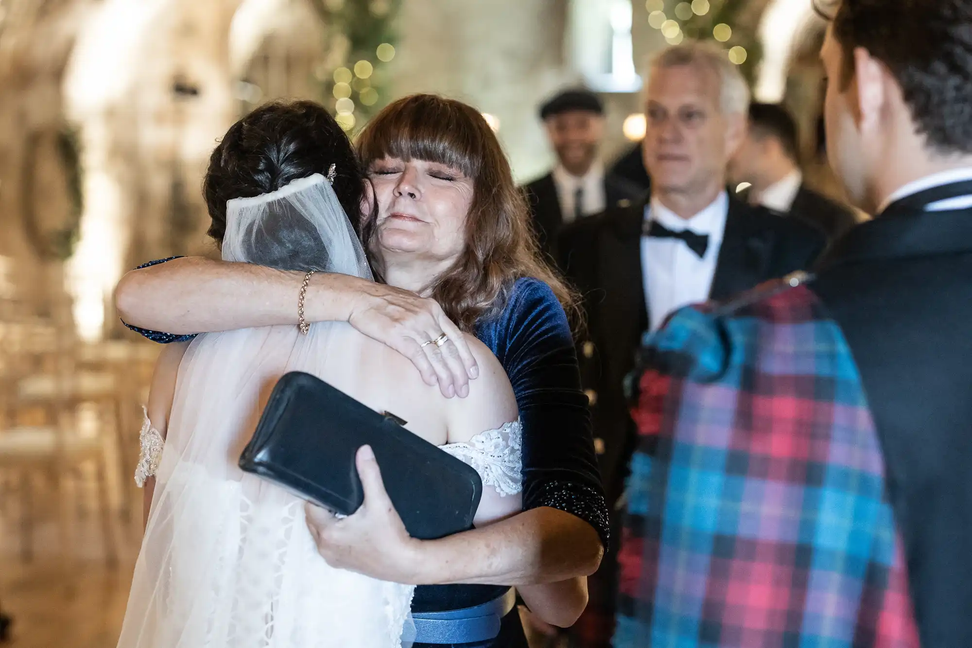 A woman with a veil hugs an older woman holding a black clutch. Two men stand in the background; one wears a black tuxedo and the other a colorful tartan outfit.