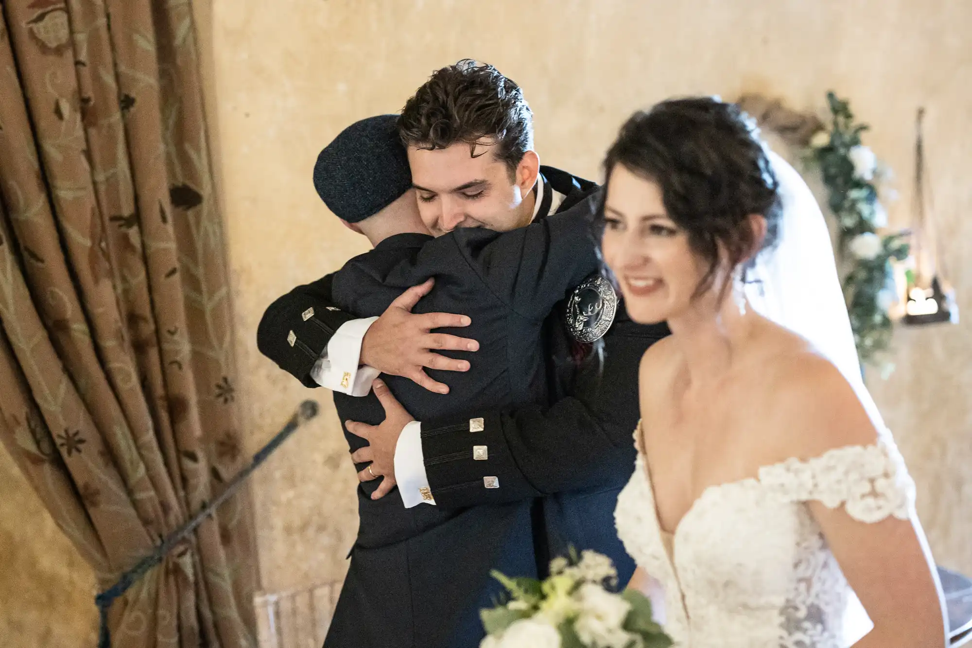 A groom hugs a man in formal attire while the bride, in a white dress, smiles and looks away. The background includes a curtain and greenery.