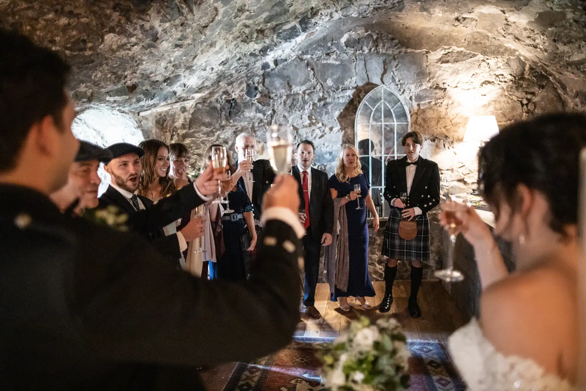 A group of people in formal attire raise their glasses for a toast in a stone-walled room with arched windows.