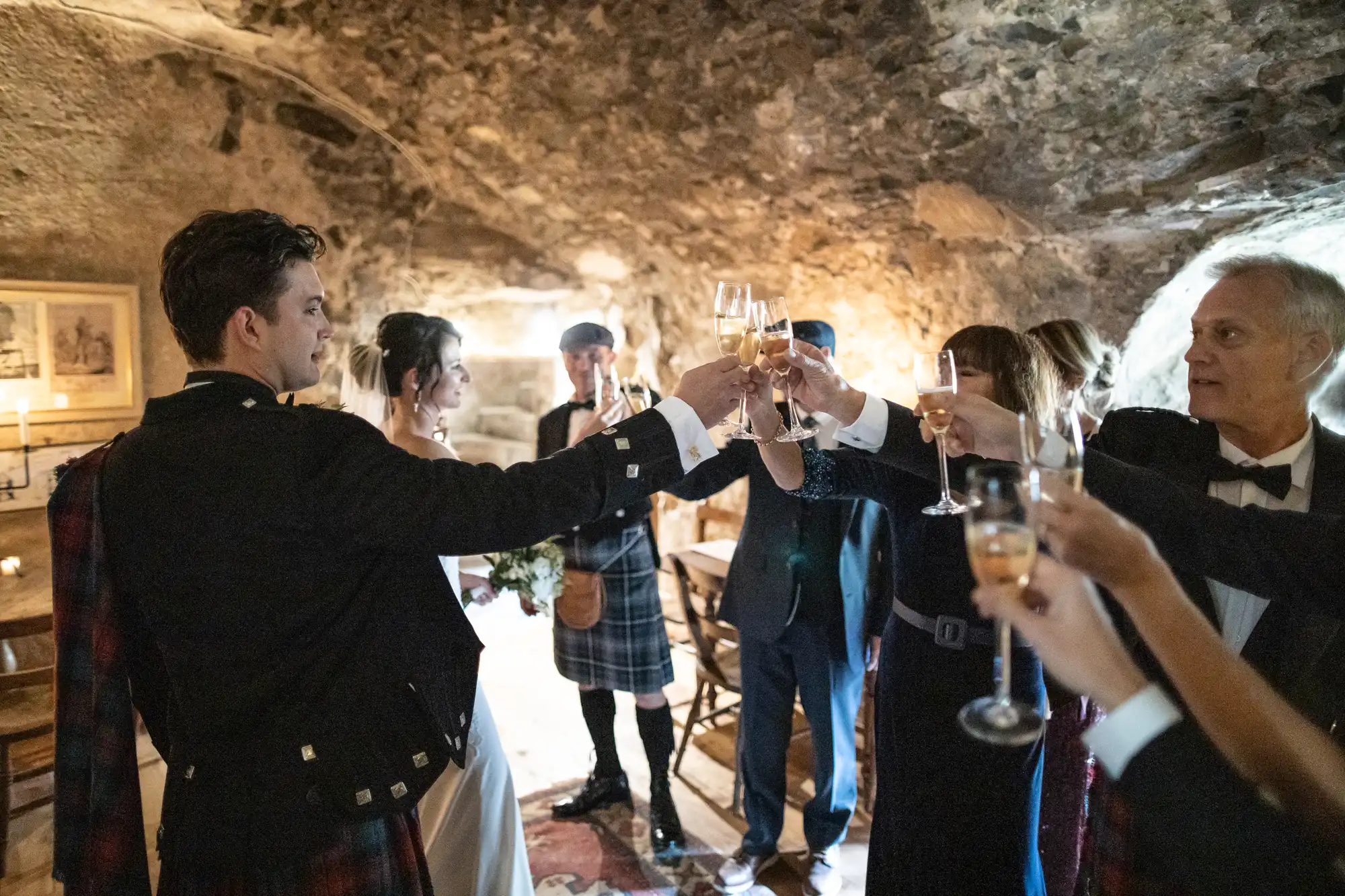 A group of people in formal attire, some wearing traditional Scottish kilts, raise their glasses in a toast inside a rustic stone-walled room.