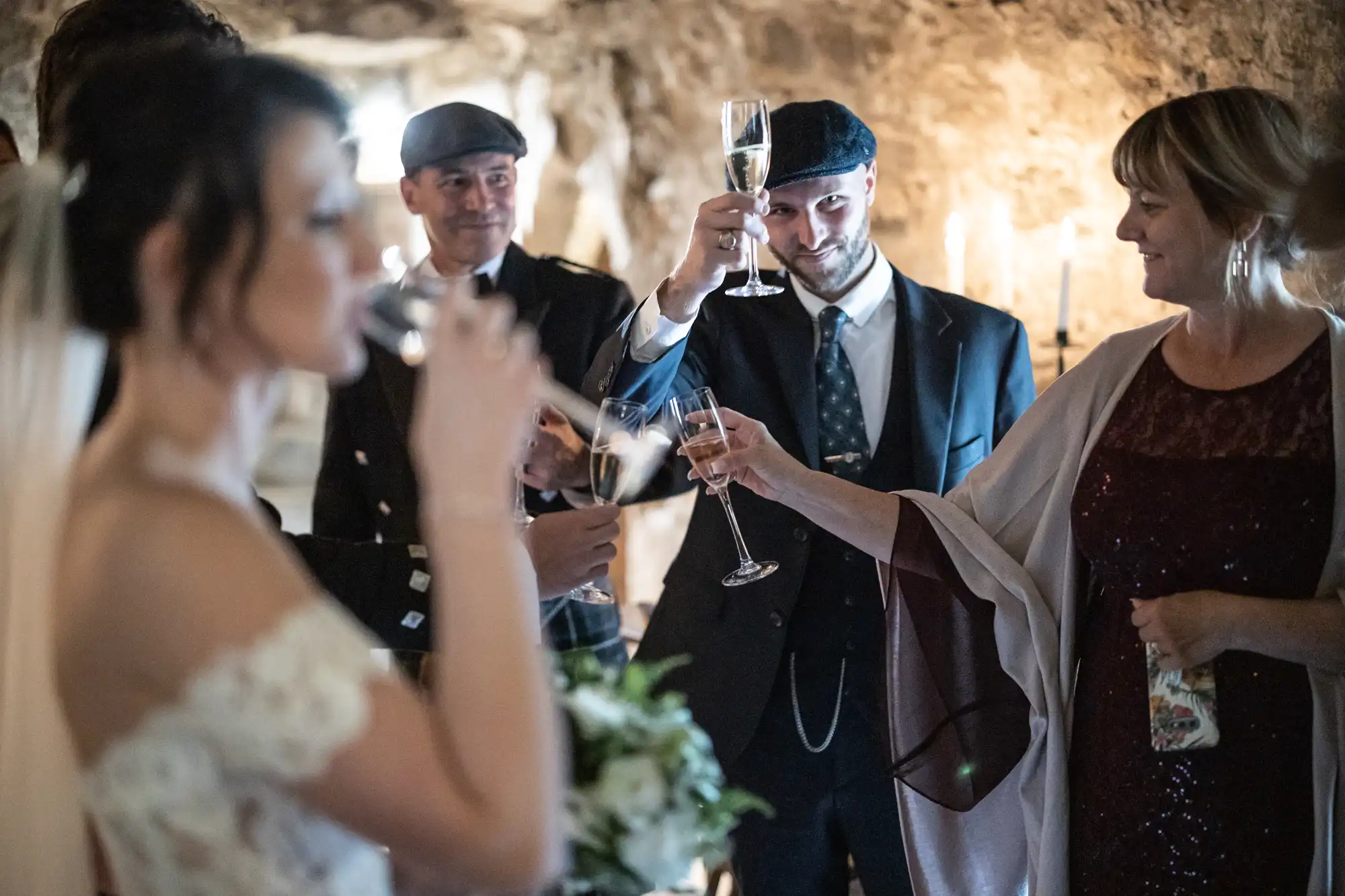 A group of people in formal attire toast with champagne glasses in a dimly lit setting. The focus is on a man and woman clinking glasses, while a bride is in the foreground drinking.