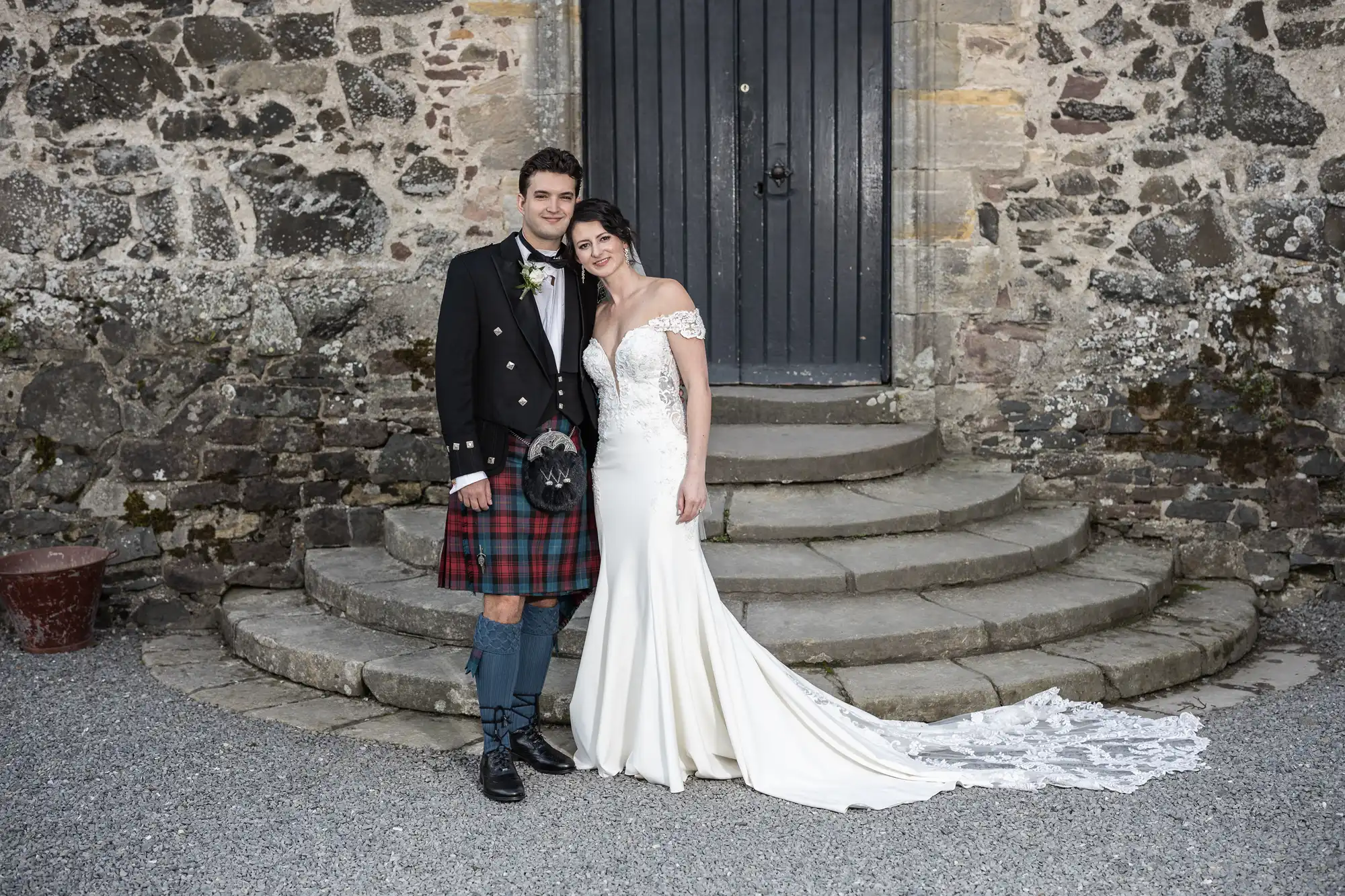 A couple in formal attire pose on stone steps in front of a rustic door. The man wears a kilt and jacket, while the woman wears a white off-the-shoulder wedding gown with a train.