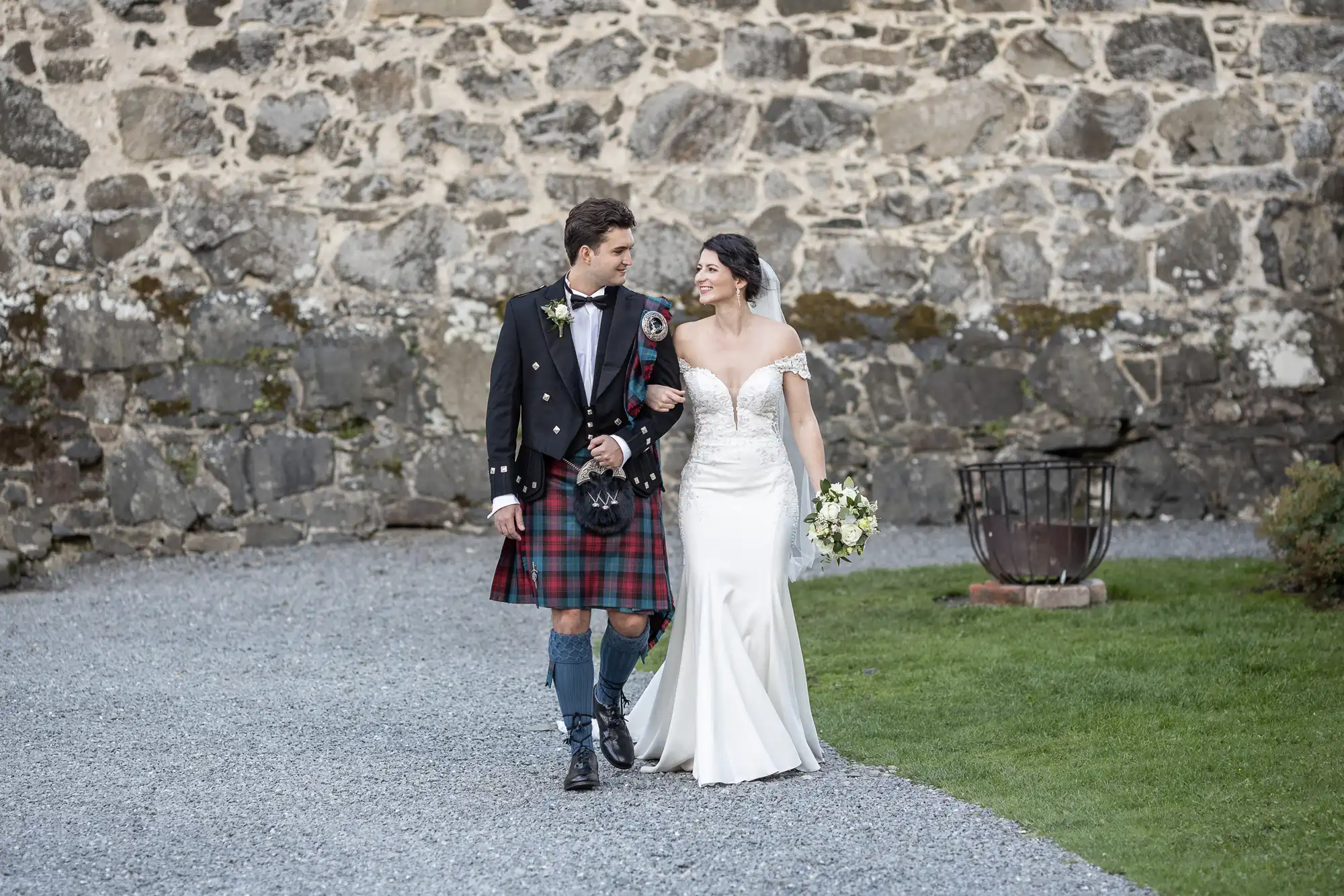 A couple in wedding attire walks arm-in-arm on a gravel path with a stone wall in the background; the groom wears a traditional Scottish kilt, and the bride wears a white gown and holds a bouquet.