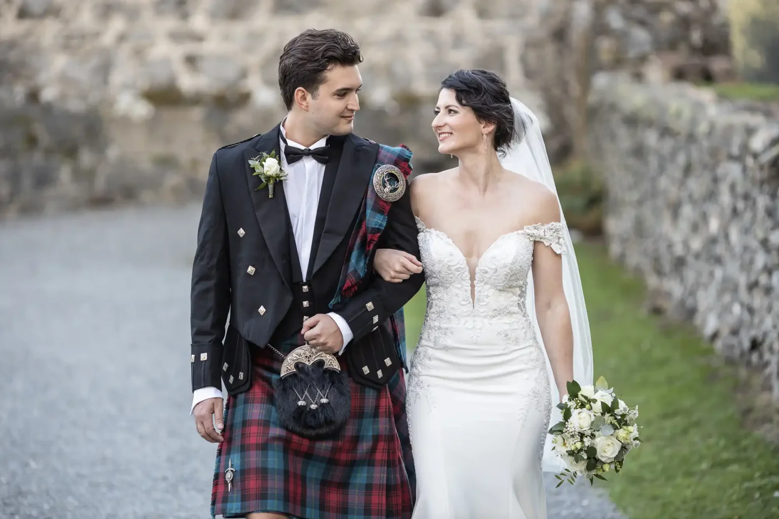 Neidpath Castle wedding photographer photo of a bride and groom walking arm in arm outdoors. The groom wears a traditional Scottish kilt, and the bride wears a white off-the-shoulder wedding dress, holding a bouquet. They both are looking at each other.