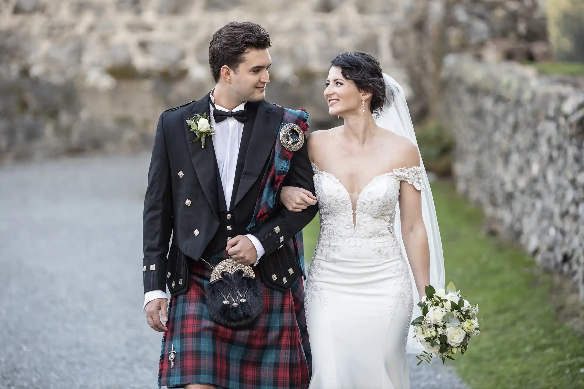 A bride and groom walk arm in arm outdoors. The groom wears a traditional Scottish kilt, and the bride wears a white off-the-shoulder wedding dress, holding a bouquet. They both are looking at each other.