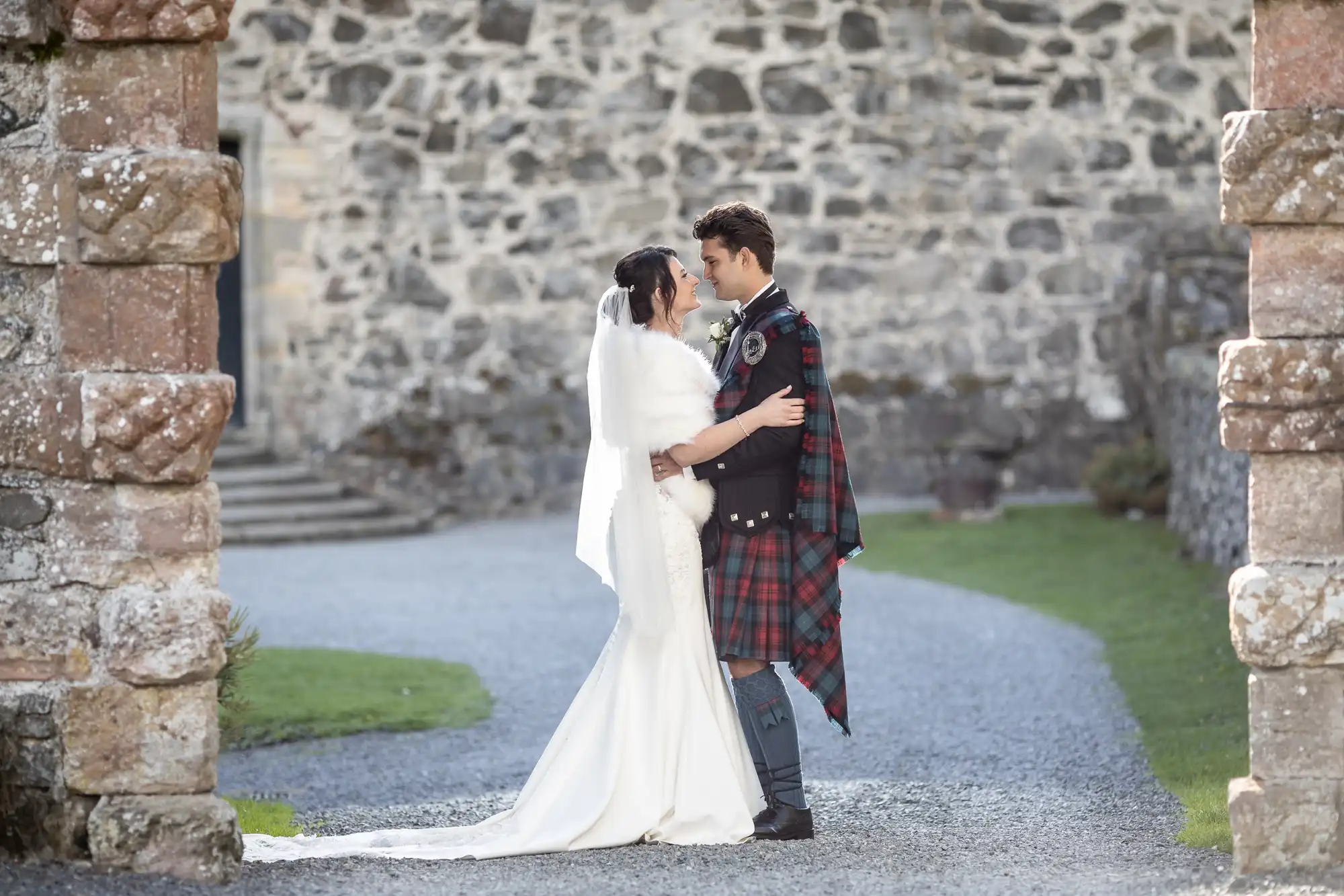 A bride in a long white gown and a groom in a traditional Scottish kilt embrace and look at each other while standing outdoors in front of a stone wall.