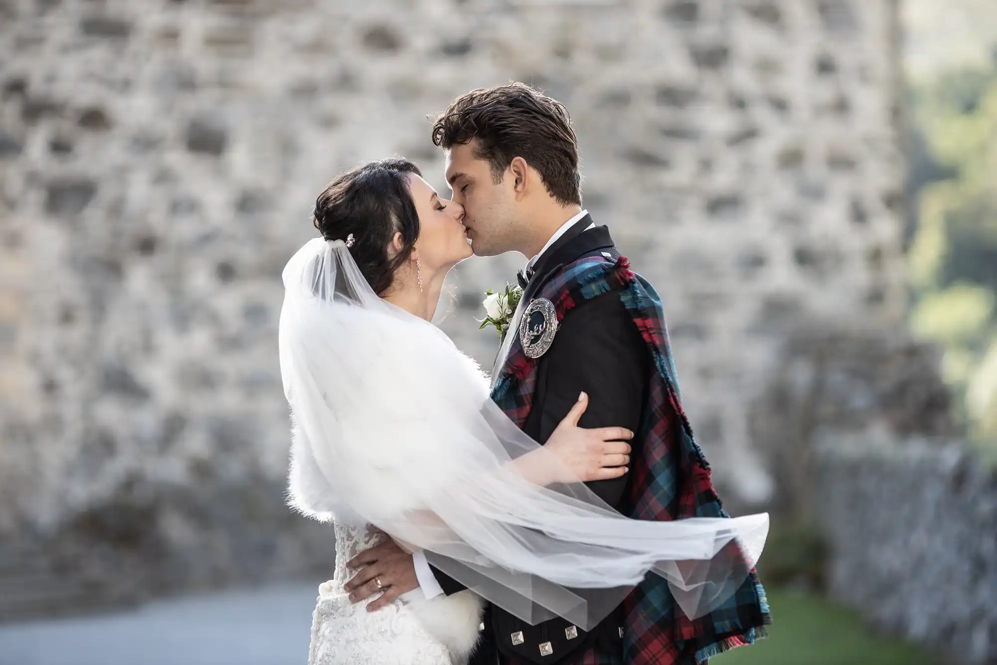 A bride and groom kiss outdoors, with the groom wearing a traditional Scottish kilt outfit and the bride in a white wedding dress and veil. A stone wall is visible in the background.