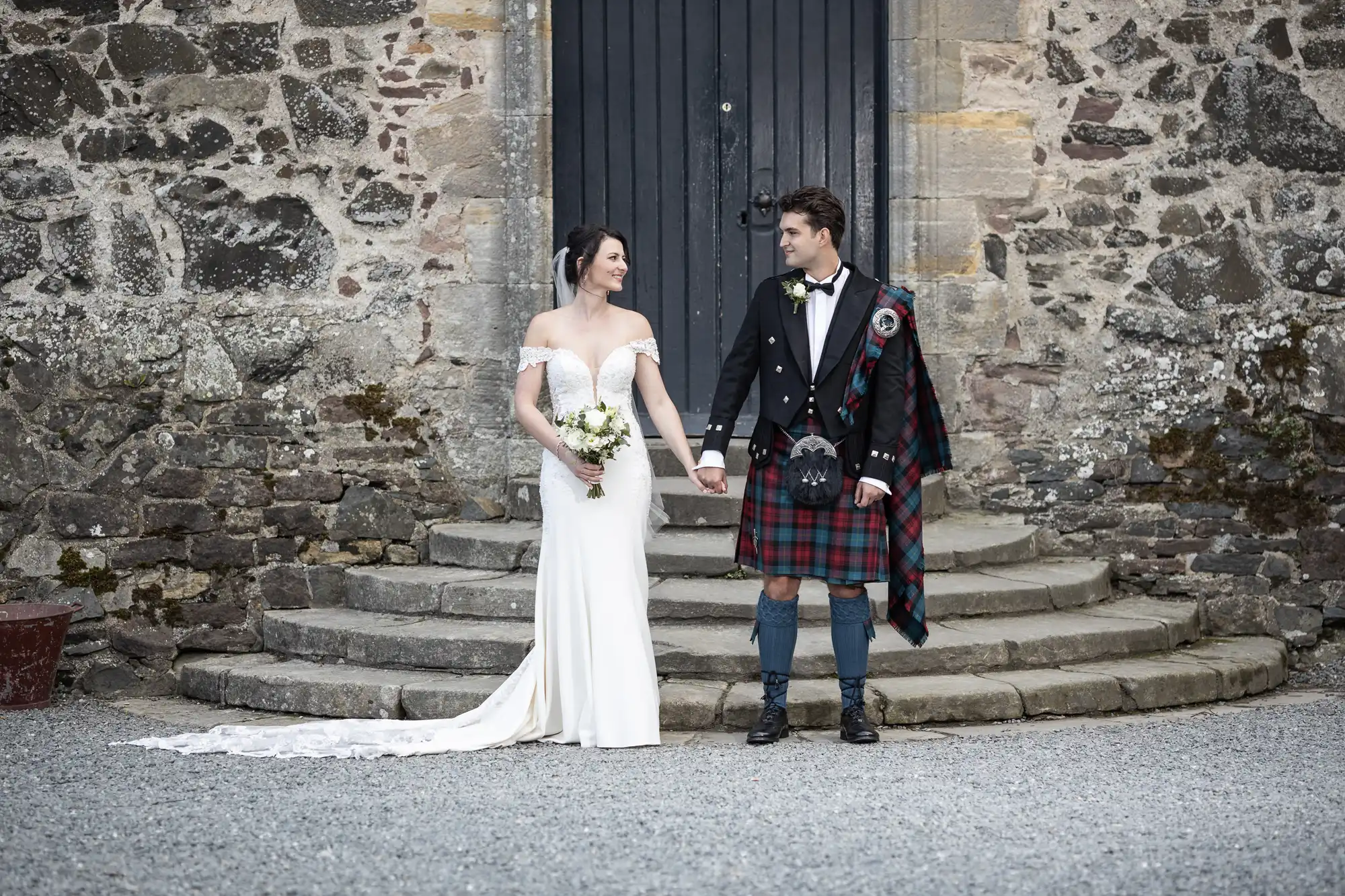 A bride in a white off-shoulder gown and a groom in a traditional Scottish kilt hold hands, standing in front of a stone building with a dark wooden door.