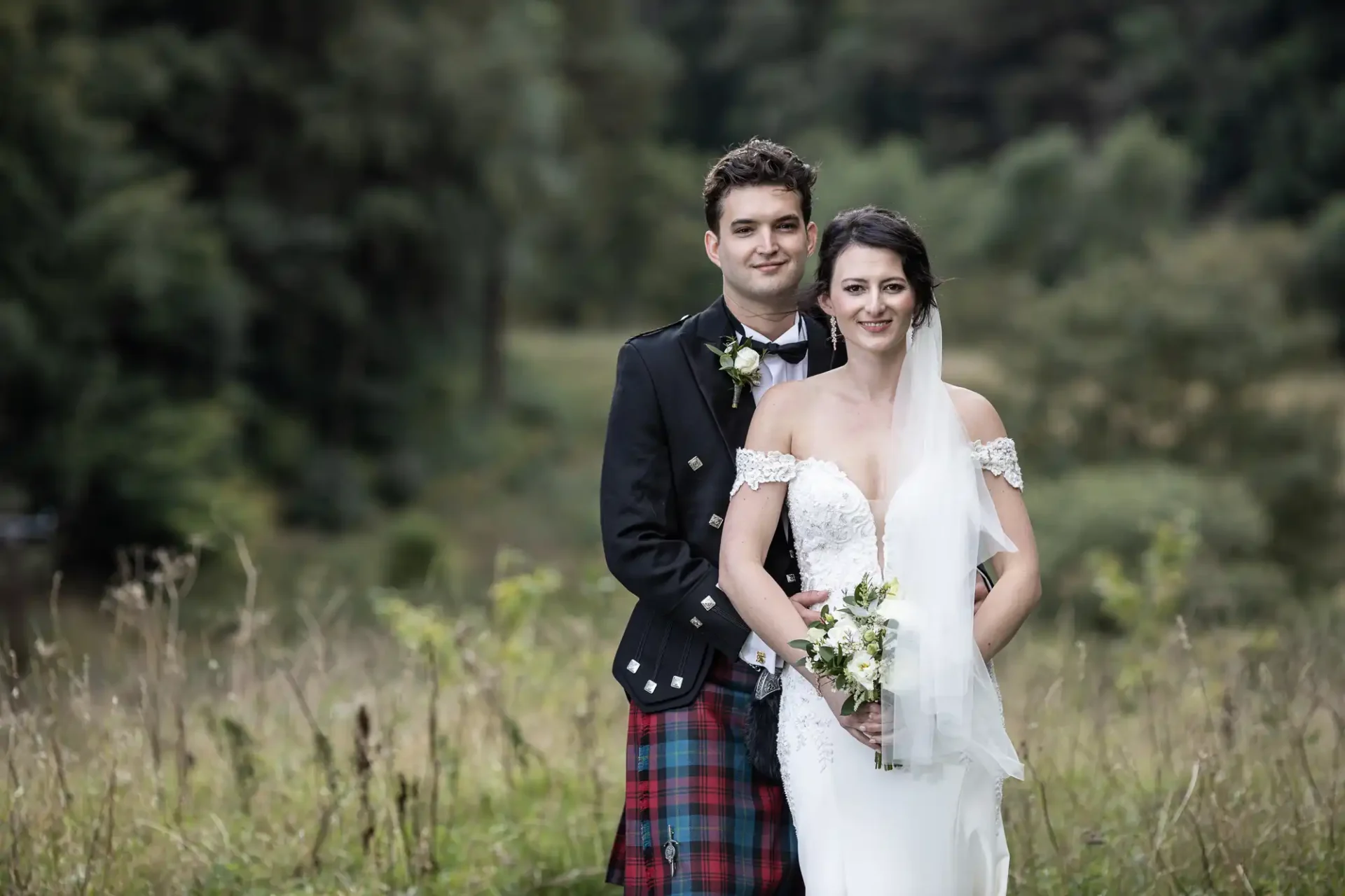 A couple stands outdoors, the man wearing a kilt and jacket, and the woman in a white wedding dress holding a bouquet. They are surrounded by greenery, with trees in the background.