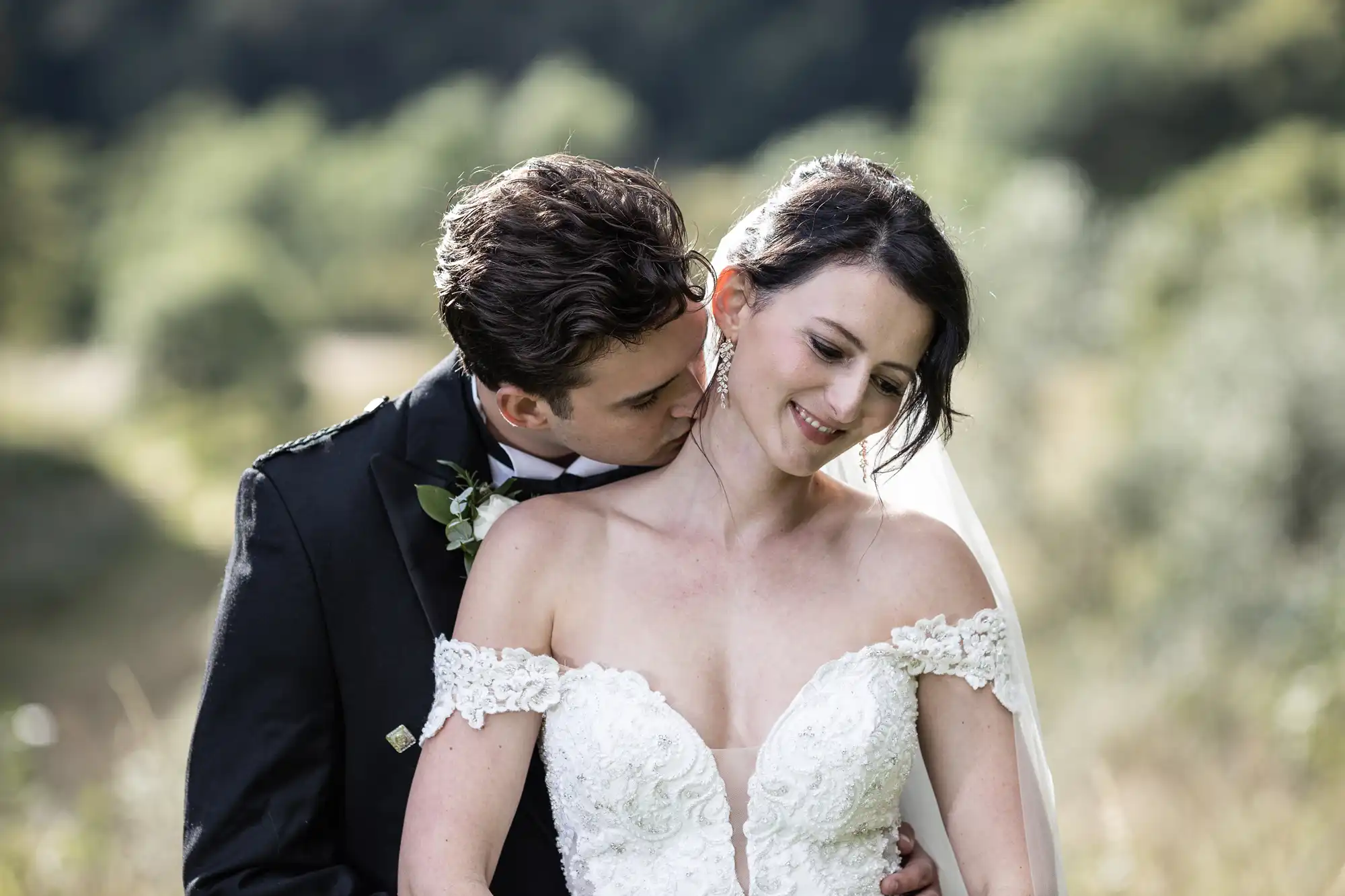 A bride and groom share a close moment outdoors. The groom hugs and kisses the bride on the neck while she smiles, eyes closed. They are in wedding attire.