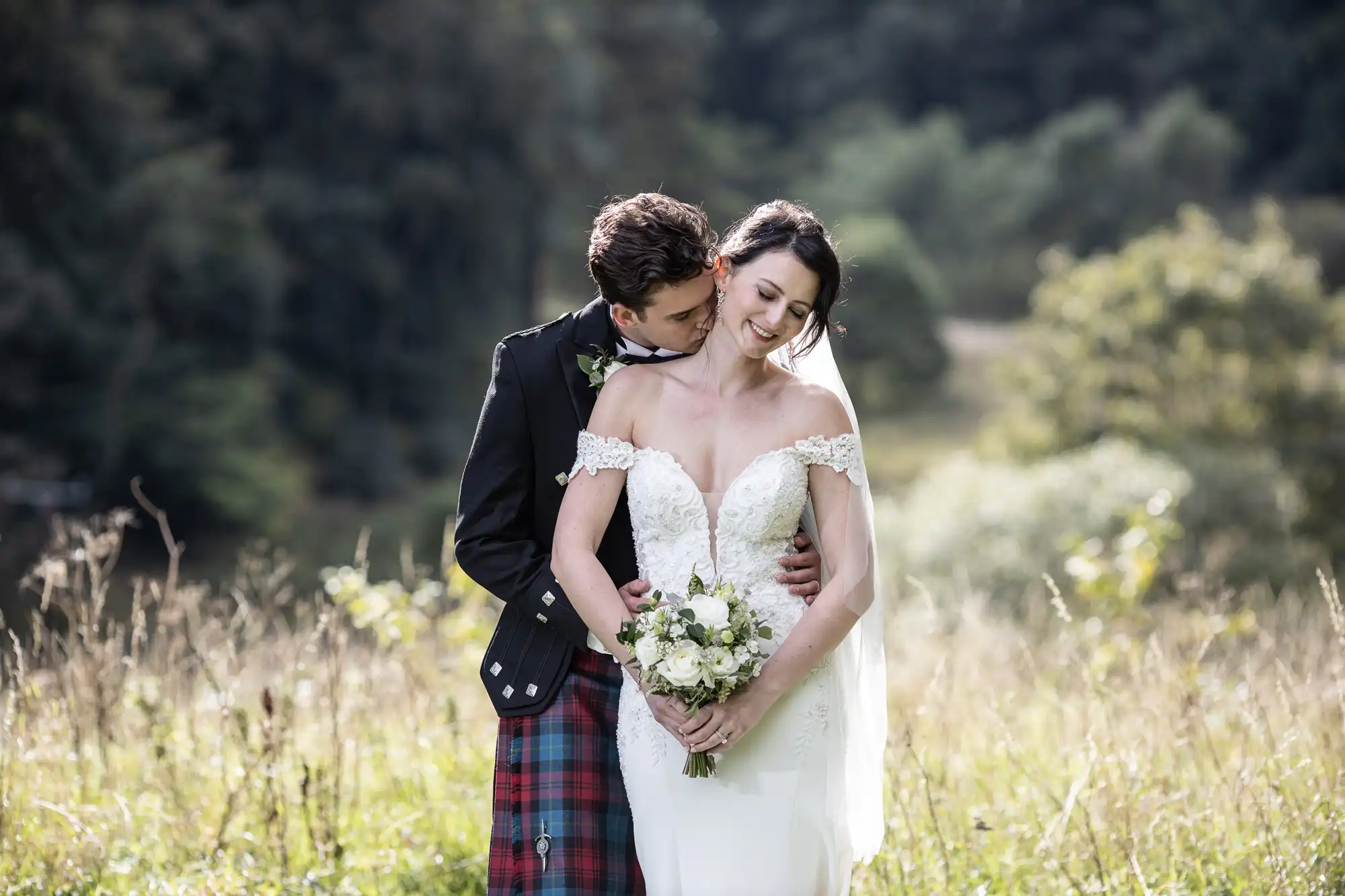 A couple stands in a field, the man embracing the woman from behind. She holds a bouquet and wears a white wedding dress, while he is dressed in a dark jacket and a red tartan kilt.