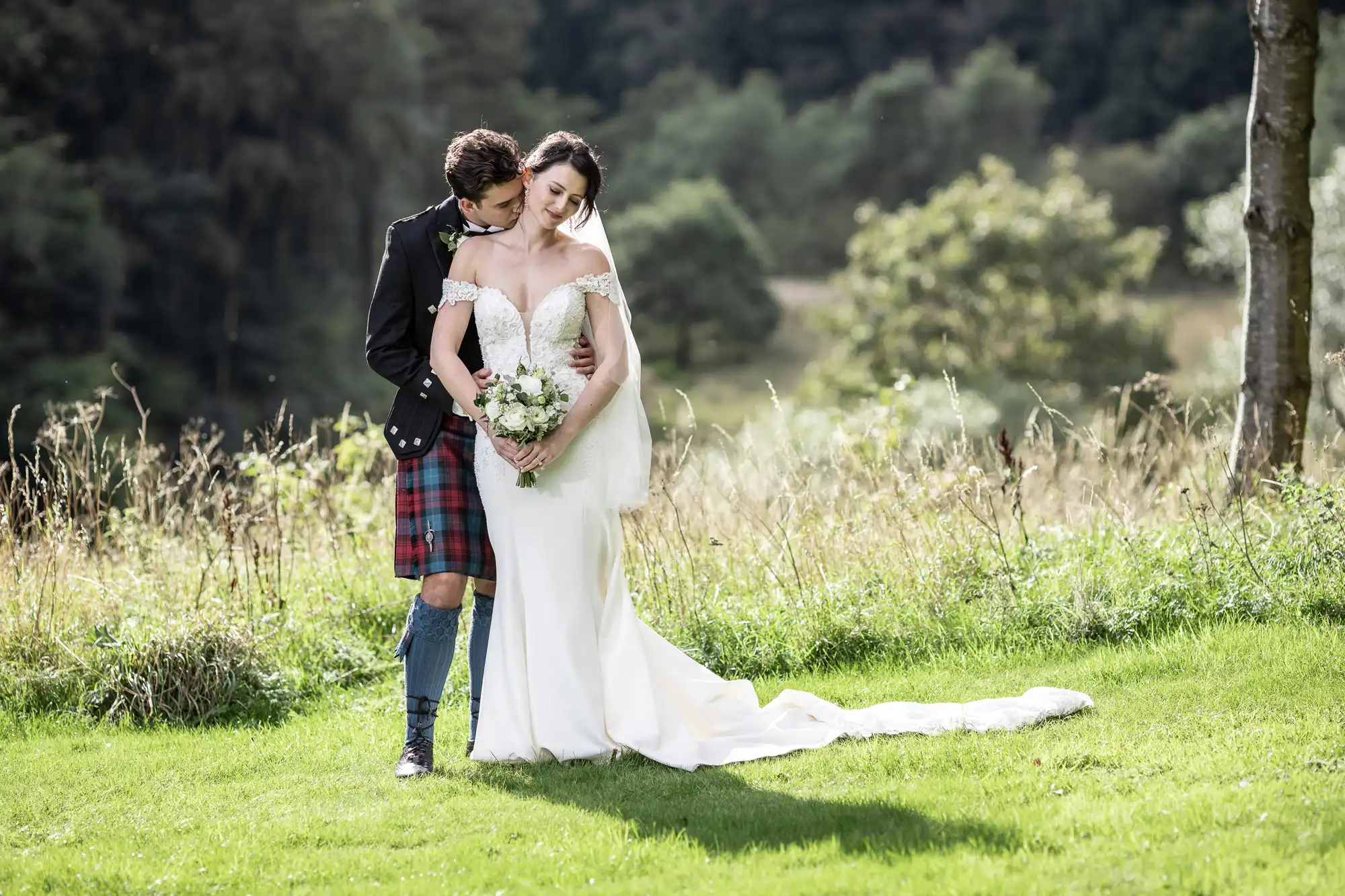 A couple stands outdoors, with the groom in traditional attire hugging the bride, who is wearing a white wedding dress and holding a bouquet. They are in a grassy area with trees in the background.