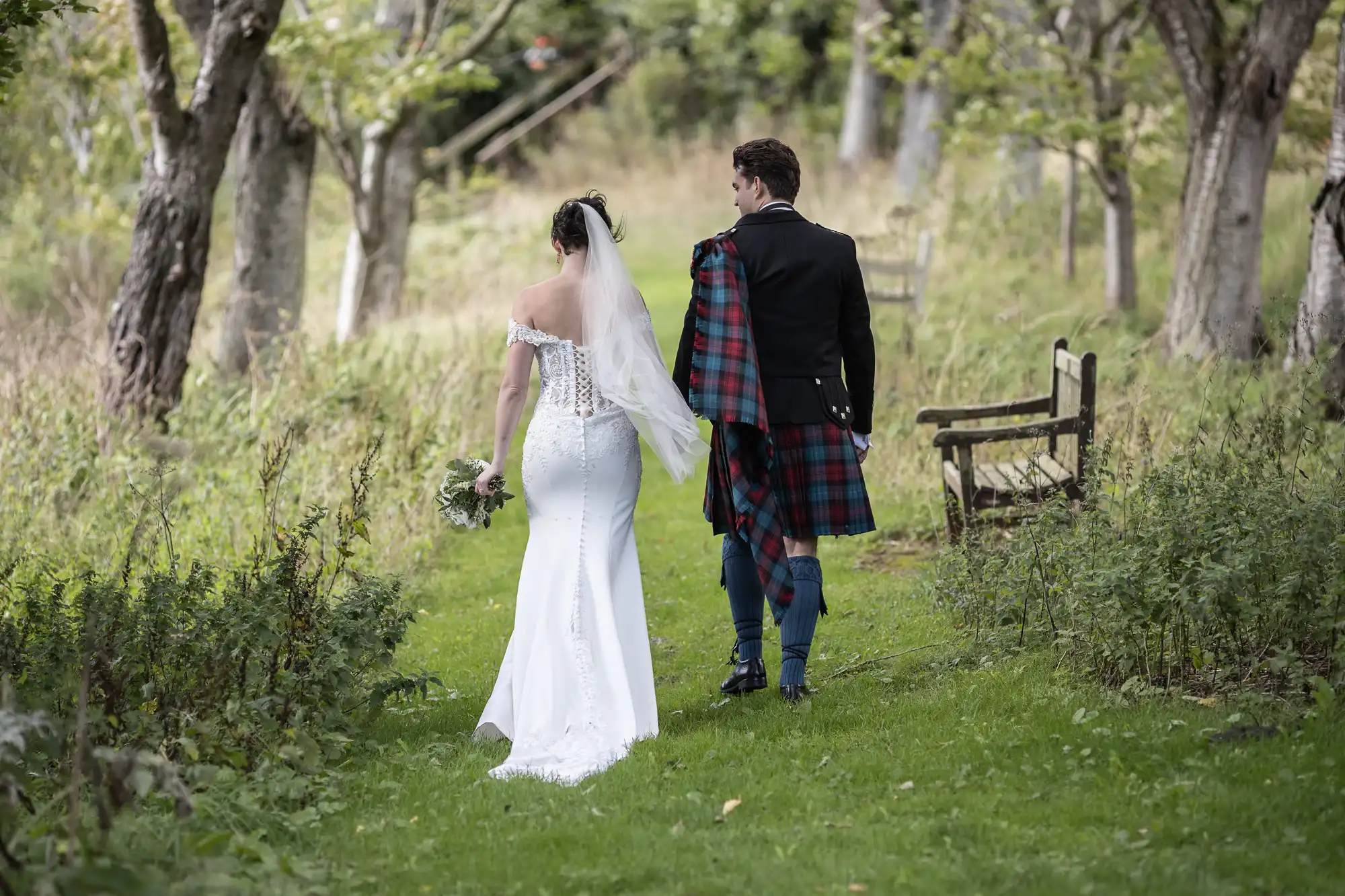 A bride and groom walk down a grassy path in an outdoor setting, viewed from behind. The bride is wearing a white dress and veil, while the groom wears a dark jacket with a plaid draped over his shoulder.