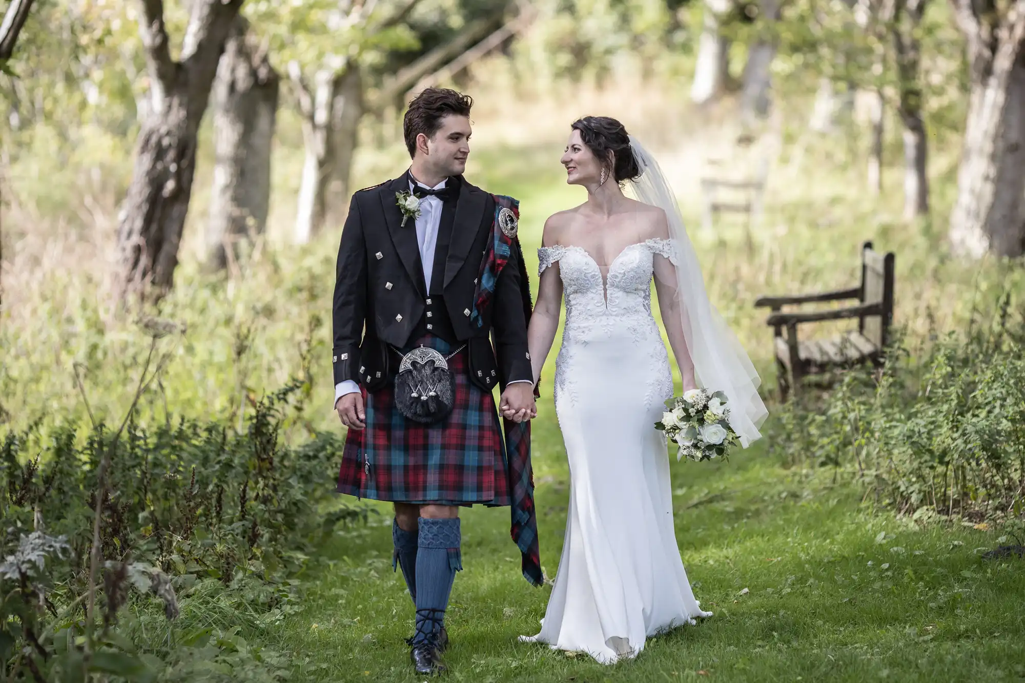 A couple walks hand in hand outdoors. The groom wears a traditional Scottish kilt and jacket, while the bride wears an off-shoulder white wedding dress and veil, holding a bouquet.