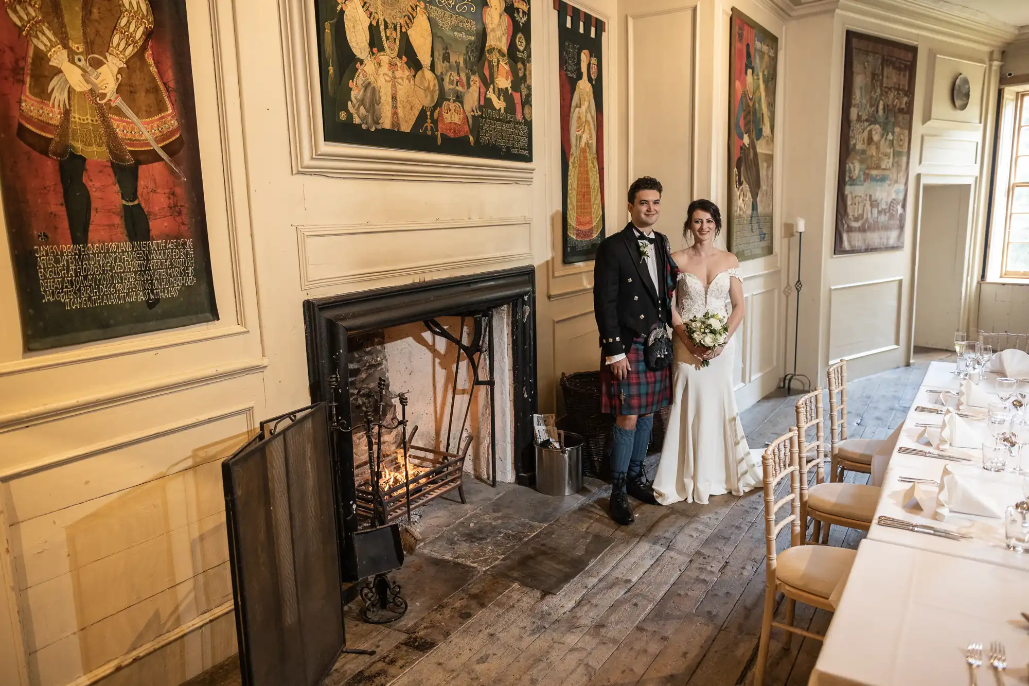 A bride and groom in wedding attire stand beside a fireplace in a historic room with decorated walls and table settings.