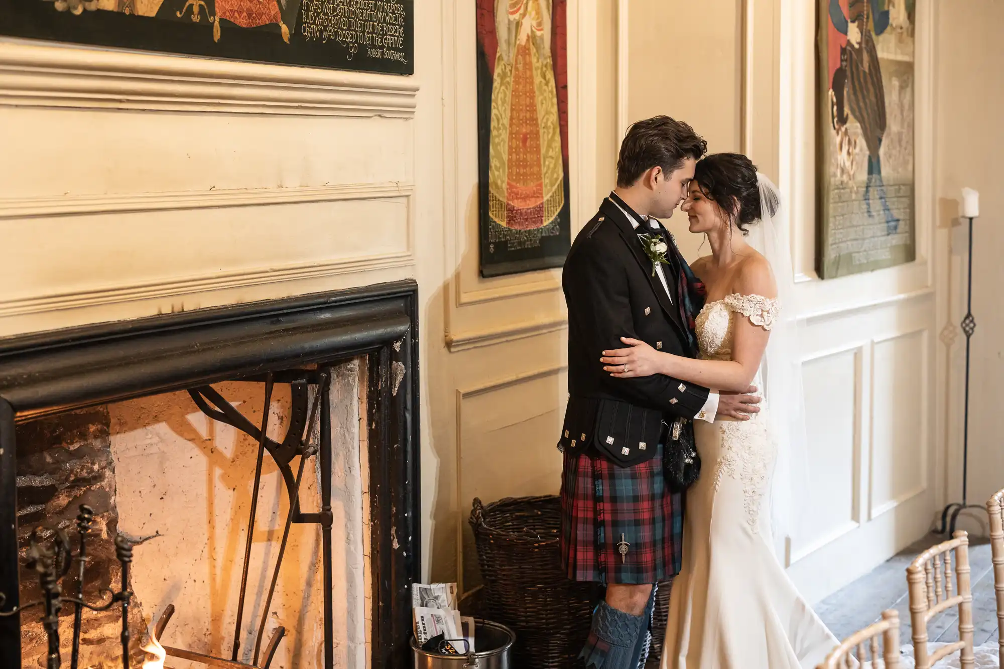 A couple stands closely, embracing lovingly next to a fireplace in a rustic room adorned with wall art. The man is wearing a kilt, and the woman is in an elegant white dress.