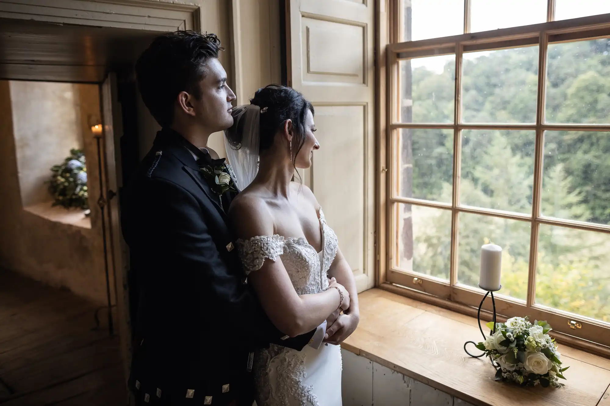 A couple dressed in a wedding gown and suit stands by a window. The bride and groom look outside while holding each other. A lit candle and flower bouquet are on the windowsill.