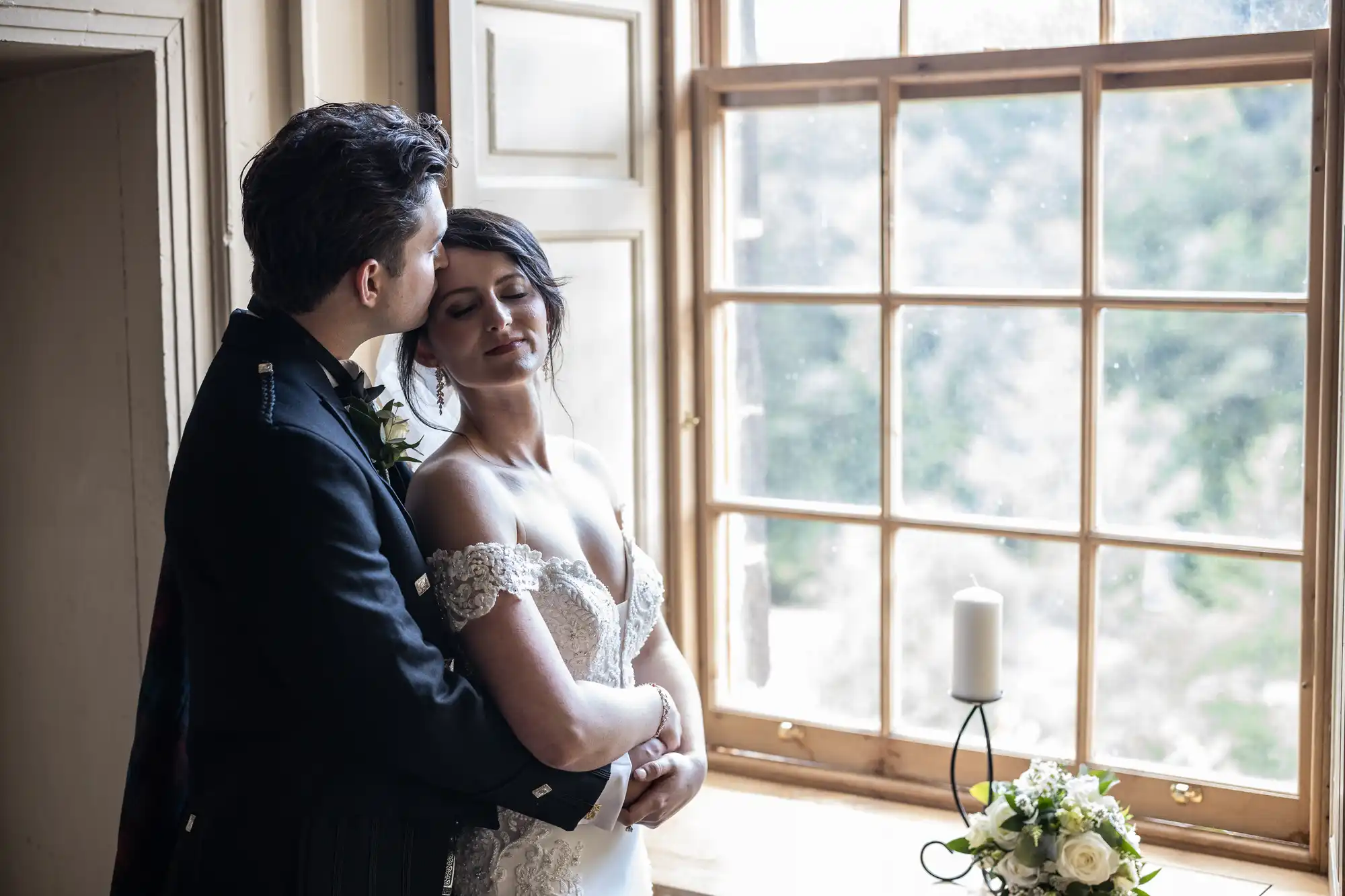 A couple dressed in wedding attire embraces tenderly by a large window with a view. A bouquet and a candle are placed on the windowsill.