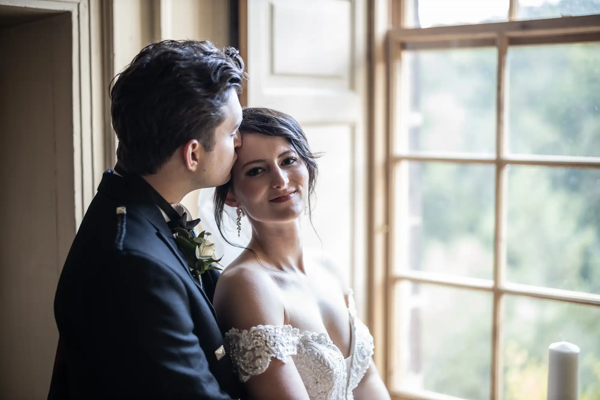 A bride and groom stand by a large window. The groom, wearing a dark suit, kisses the bride, wearing a white off-shoulder dress, on the forehead as they embrace.