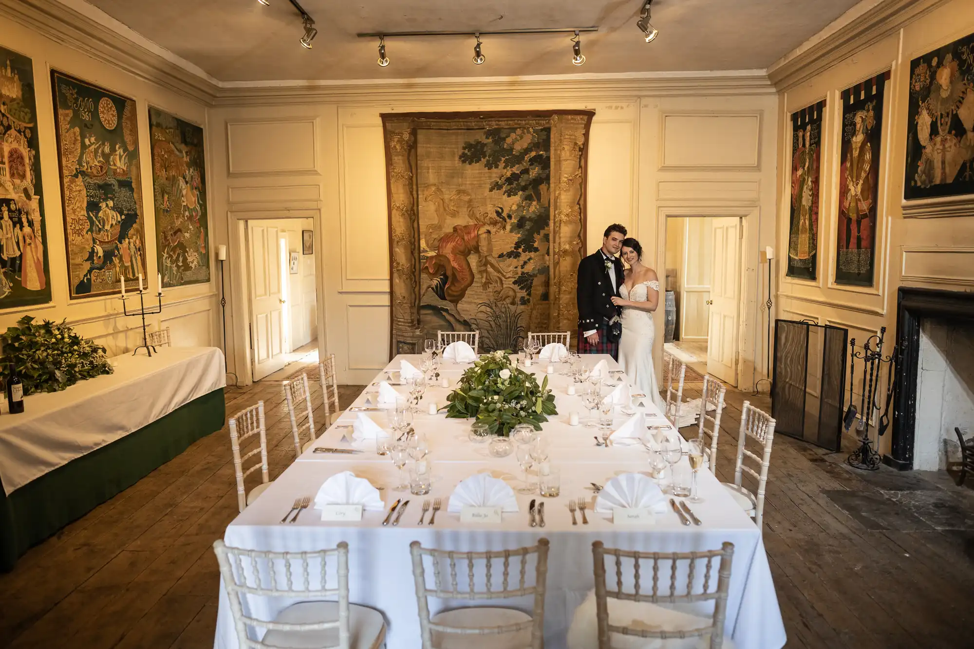 A couple in wedding attire stands behind a set dining table in an elegantly decorated room with wall tapestries and white chairs.