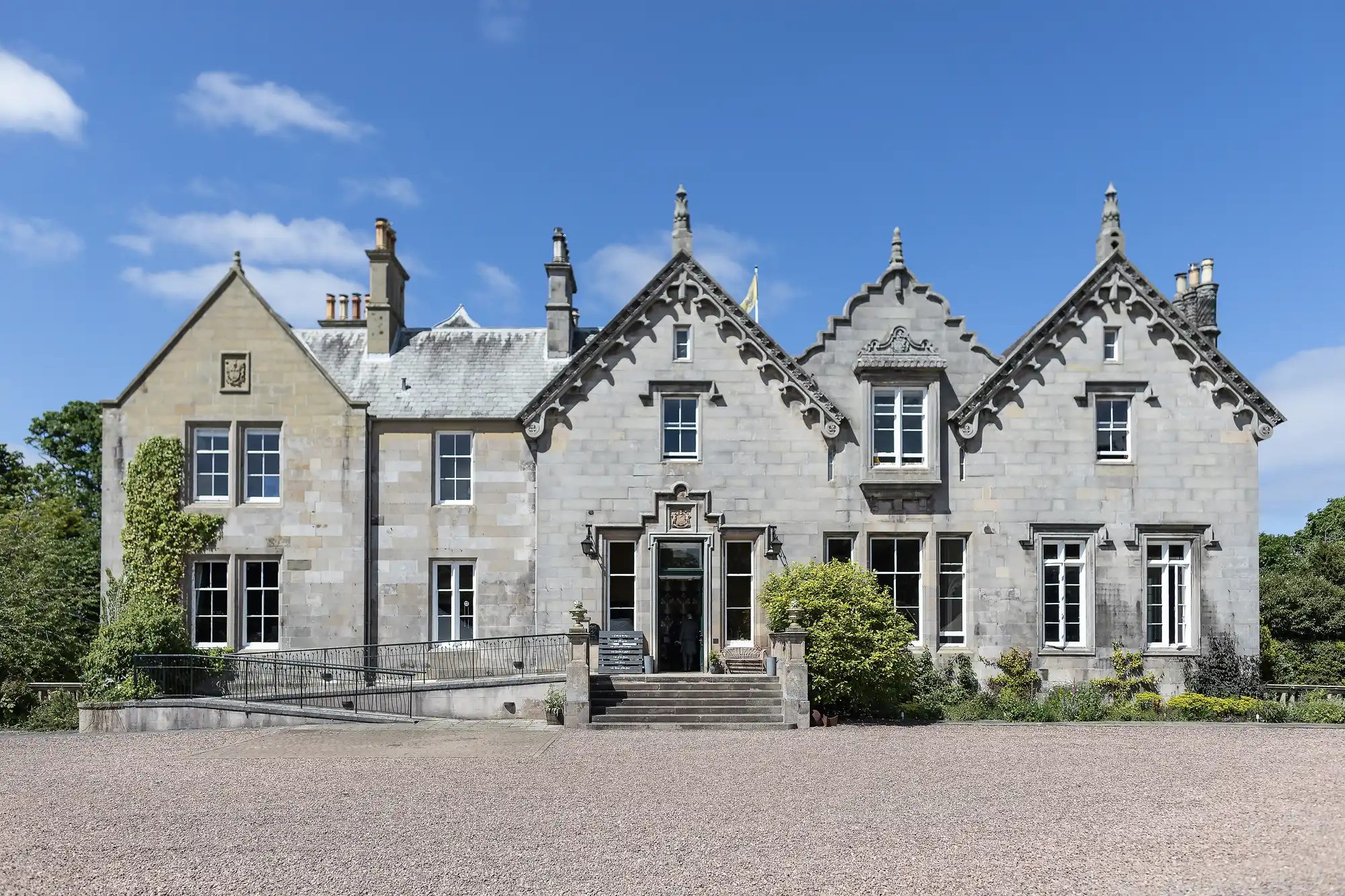 A stone mansion with multiple gables, large windows, and a central entrance. The mansion has a gravel driveway, greenery, and a clear blue sky in the background.