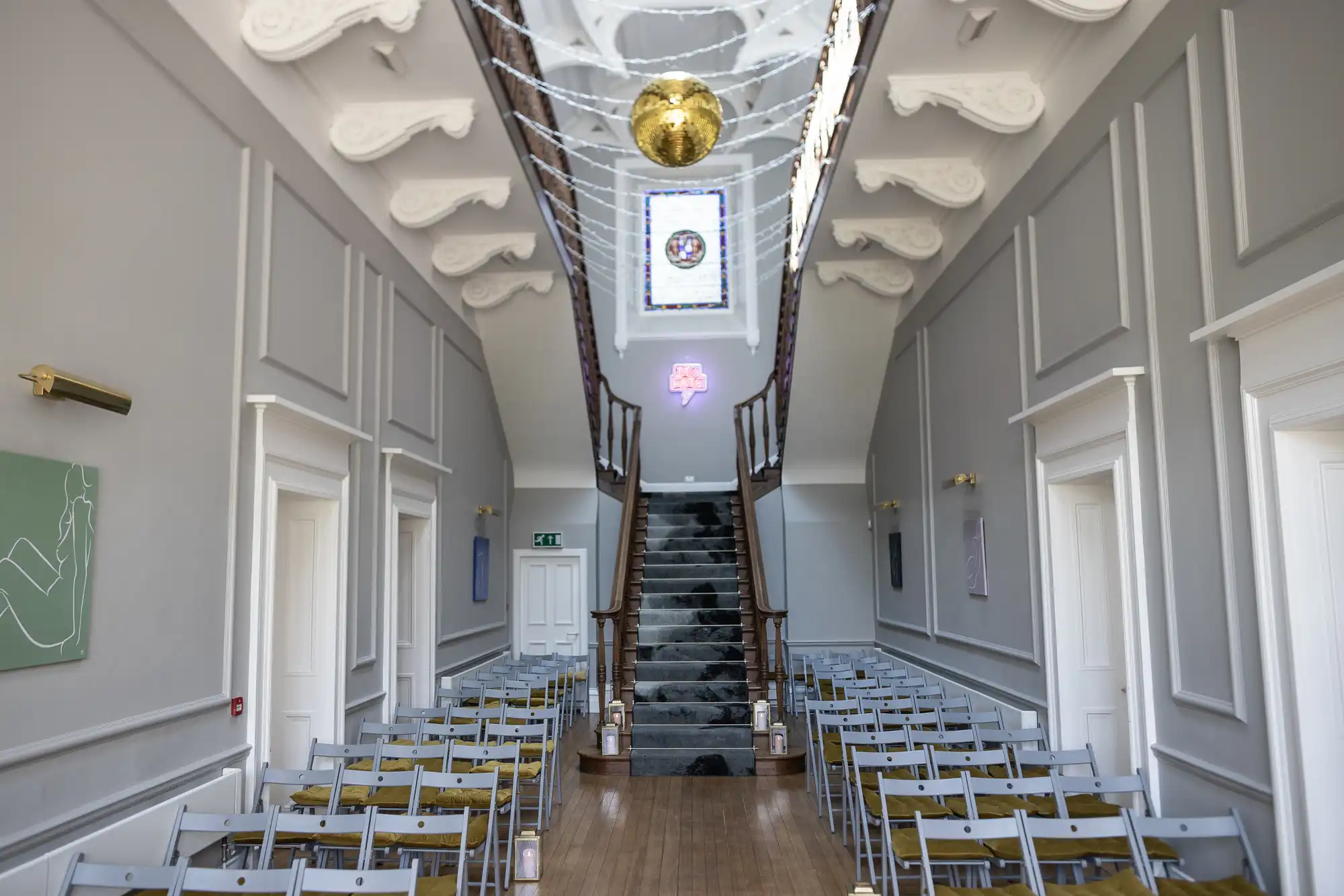 A modern indoor venue with grey folding chairs arranged in rows facing a staircase leading to a second floor. A decorative ceiling and a hanging gold sphere are visible above the staircase.