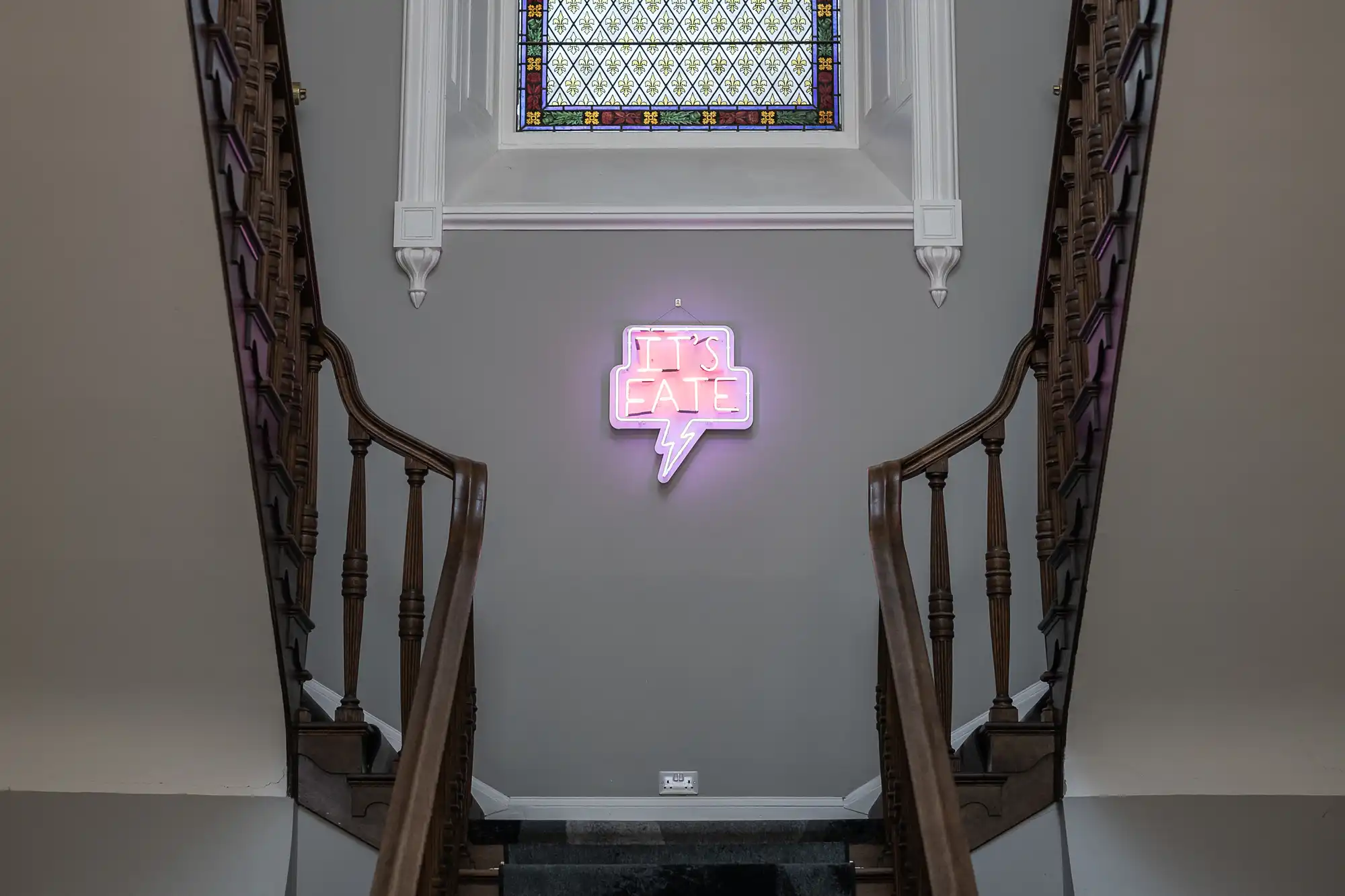 A staircase with wooden railings leads up to a wall where a neon sign that reads "IT'S LATE" is displayed, below a stained glass window.