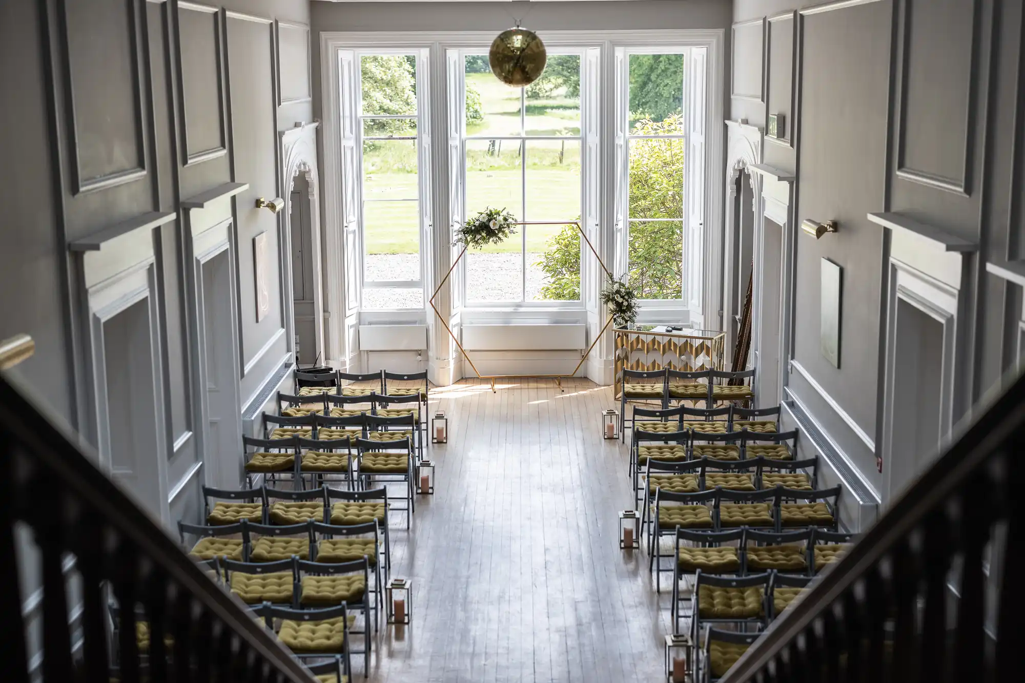 A ceremony room with rows of chairs facing a geometric wedding arch near large windows that overlook a green lawn.