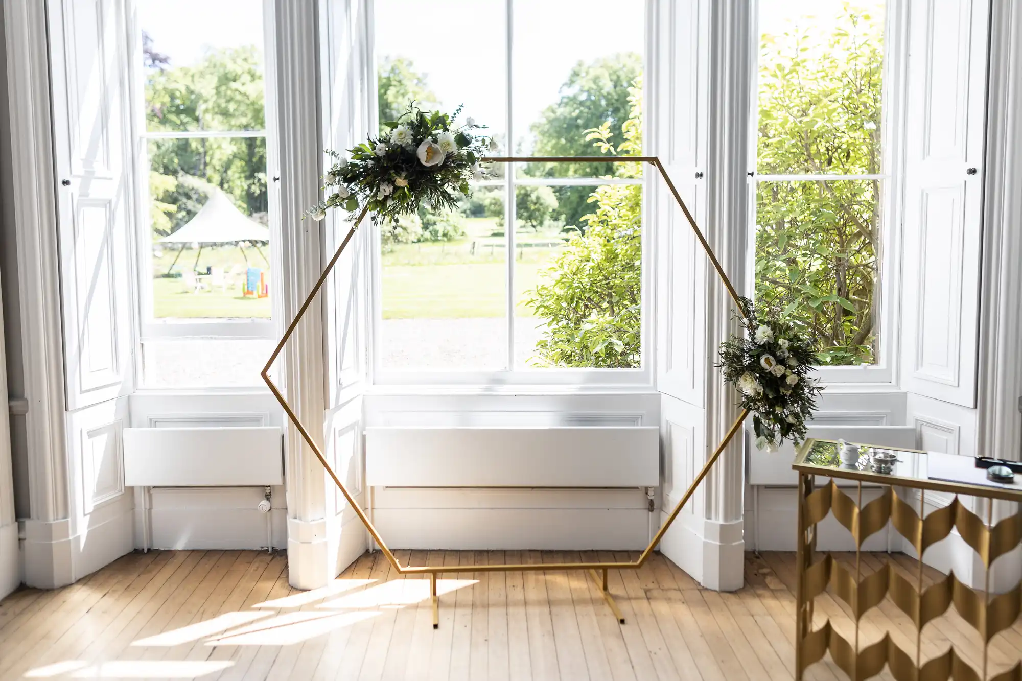 A gold hexagonal arch adorned with floral arrangements stands in front of large windows with outdoor greenery visible in the background. The room has light wooden floors.