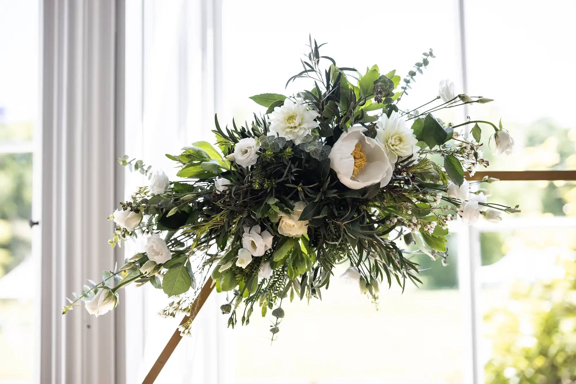 A floral arrangement featuring white flowers and green foliage is displayed indoors on a wooden stand.