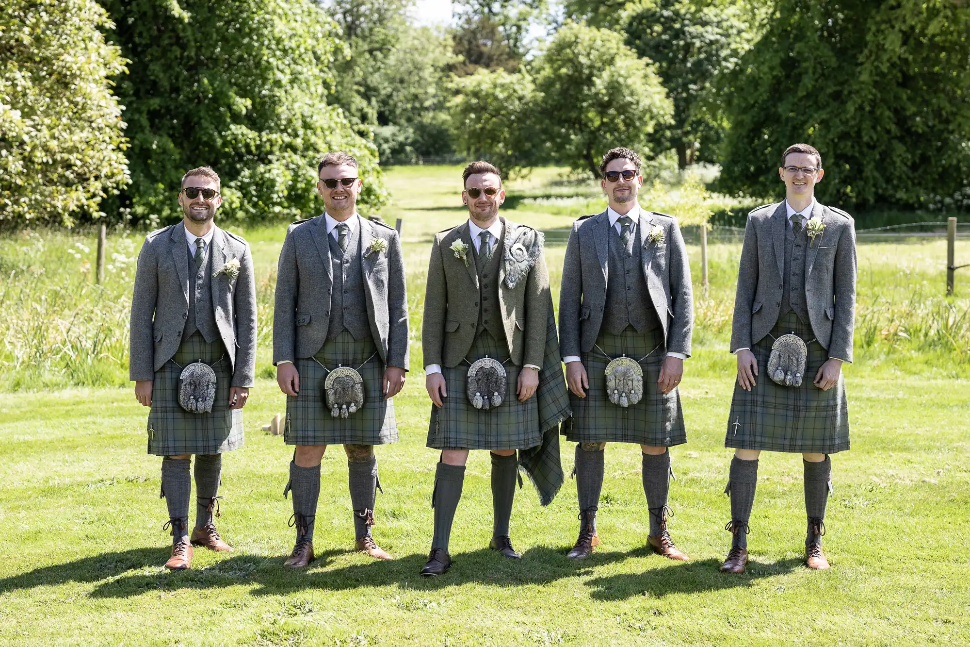 Five men wearing grey jackets and kilts stand on grass with trees in the background. They appear to be at an outdoor event.