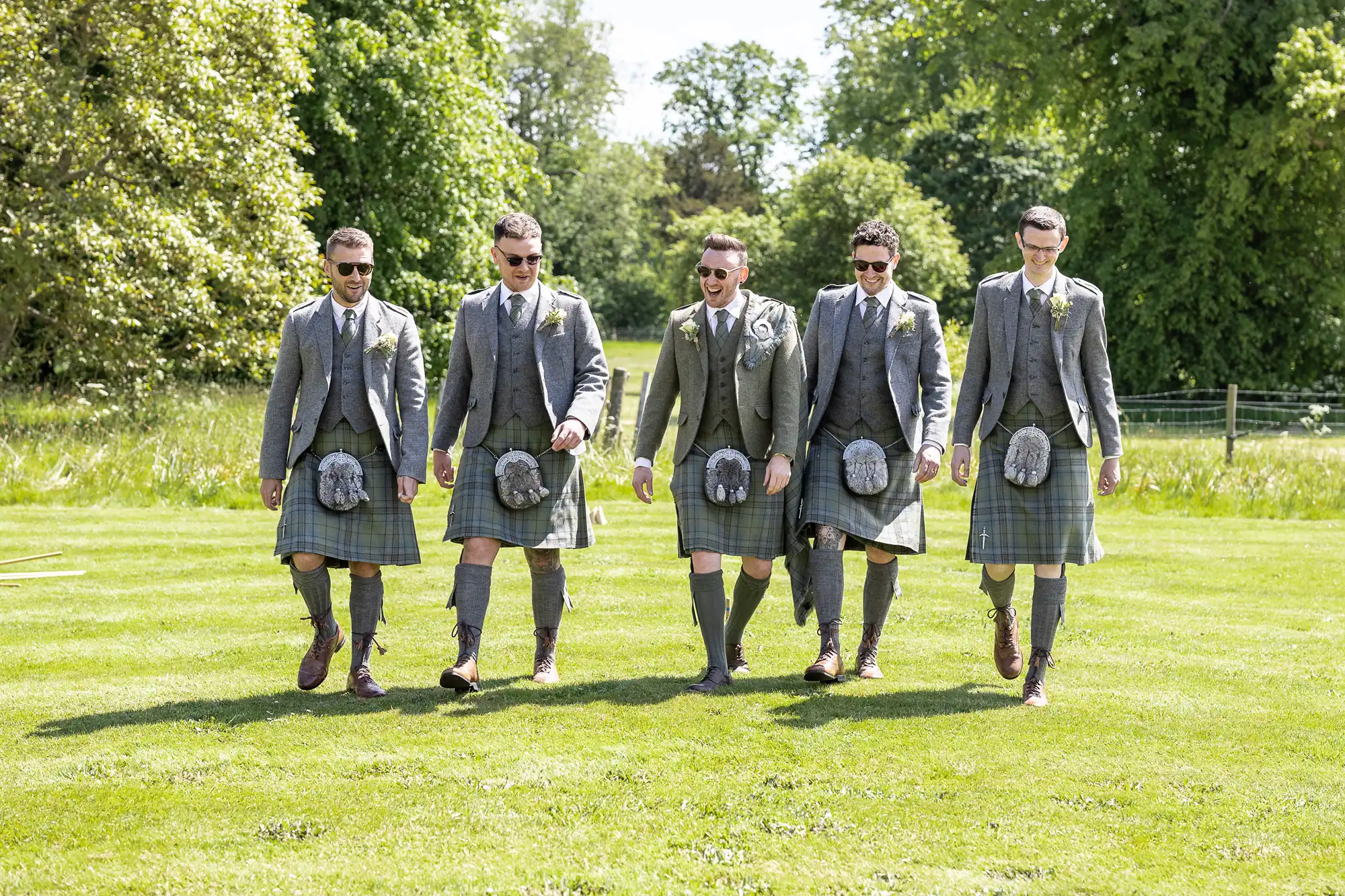 Five men wearing kilts and grey jackets walk together on a grassy lawn with trees in the background.