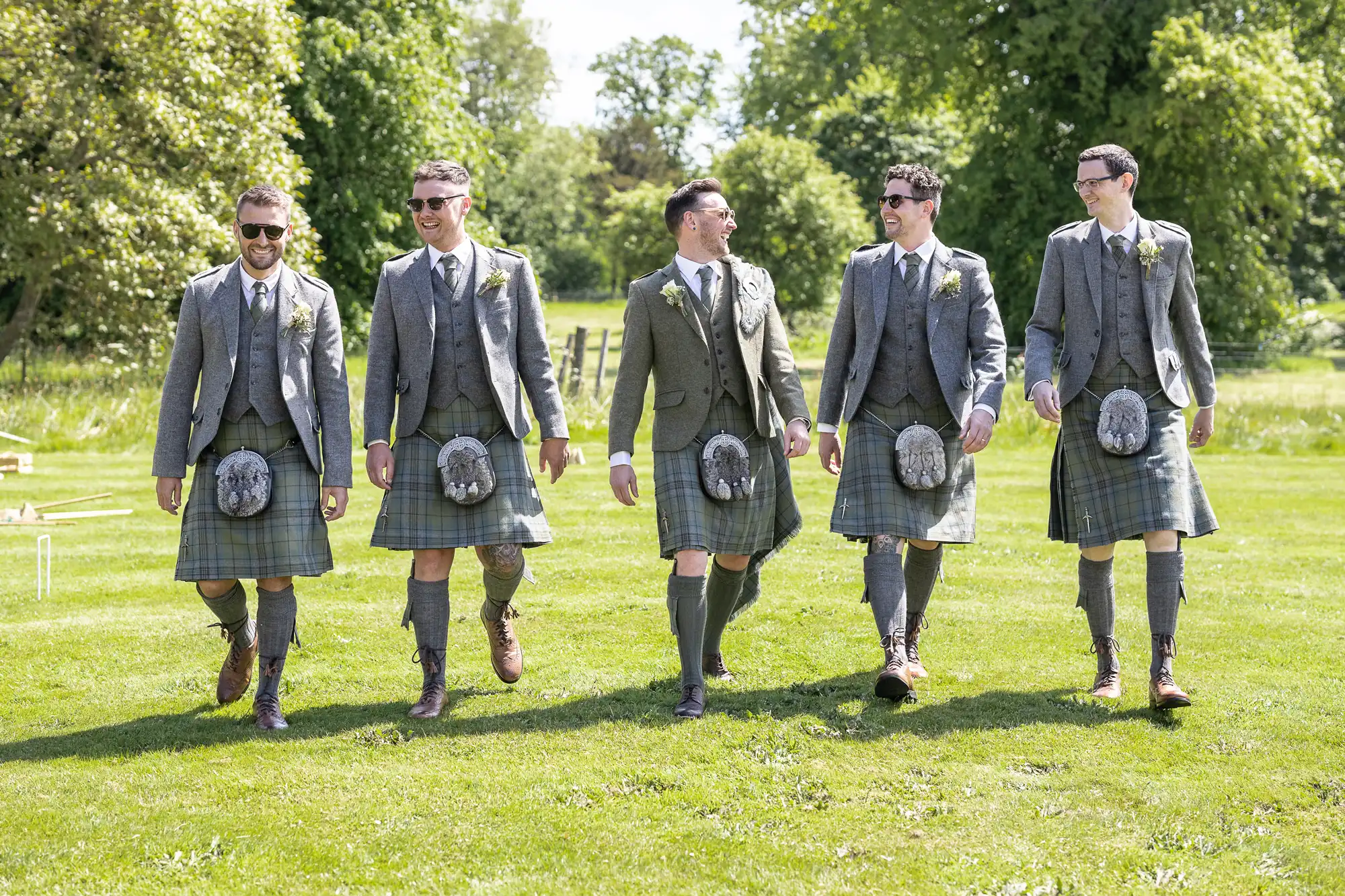 Five men in grey jackets, green kilts, and boots walk on a grassy area, smiling and engaging with each other. Trees and greenery are visible in the background.