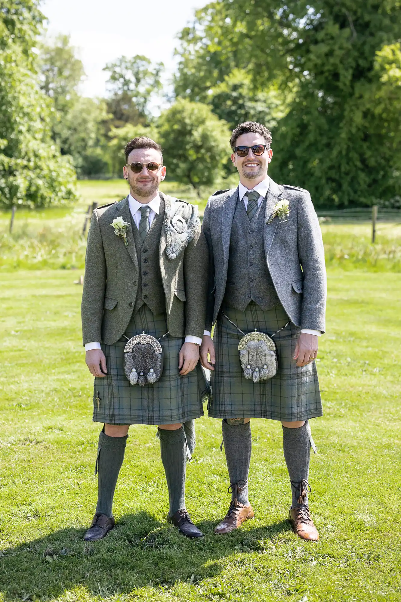 Two men stand on grass wearing traditional Scottish attire including kilts, jackets, sporrans, and sunglasses, with trees visible in the background.